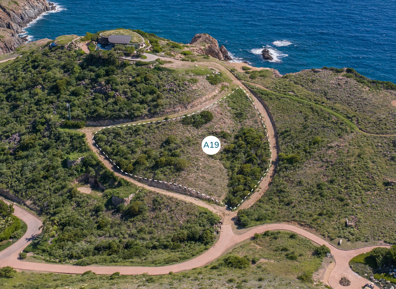 Aerial view of a grassy, hilly landscape near the ocean. A winding road with pathways and sparse vegetation leads to a circular structure atop a hill. A white marker labeled "Homesite A19" is visible in the center of the image. Waves crash against the rocky shoreline of the Atlantic Ridge.