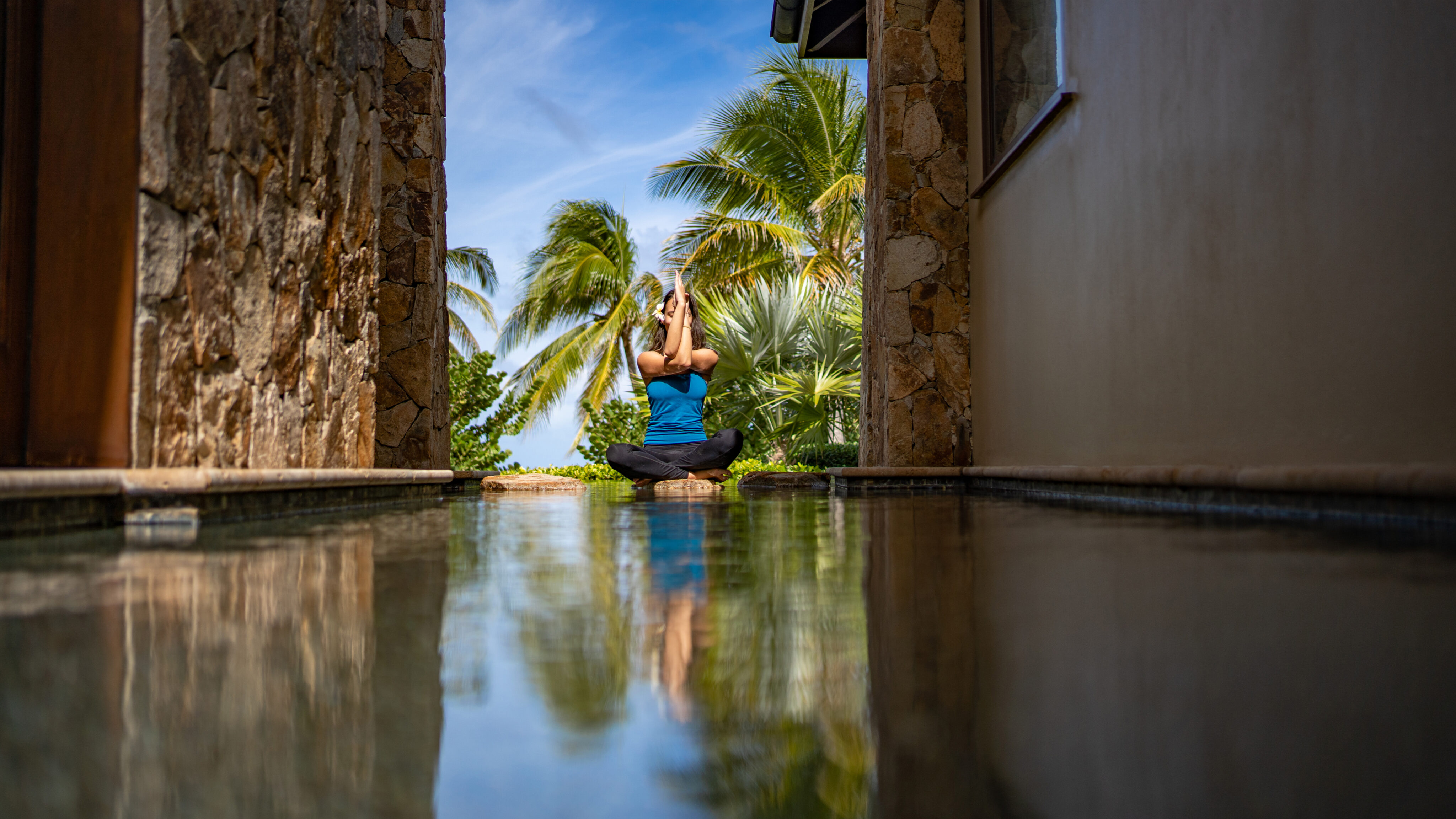 A person sits cross-legged while performing a yoga pose with hands raised above their head, surrounded by stone walls and palm trees in the background. The scene is reflected in a shallow pool of water in the foreground. Practicing yoga outdoors is among the best things to do in the BVI, especially on a bright, clear day.