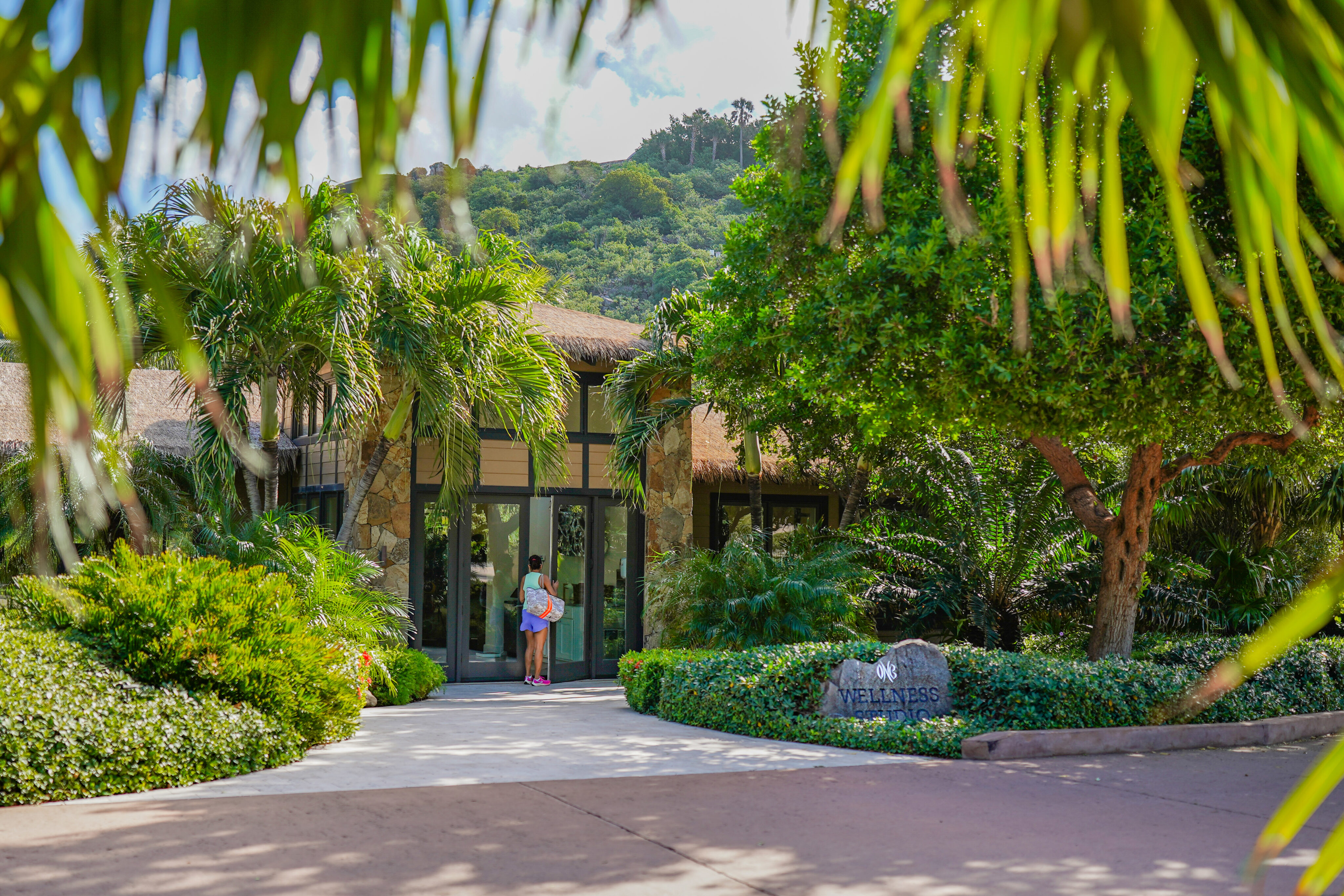 A person holding a child stands at the entrance of a building surrounded by lush tropical greenery, one of the best things to do in the BVI. Palm fronds frame the foreground, and a mountainous landscape is visible in the background. A stone sign near the entrance reads "9/12 Vitemers.