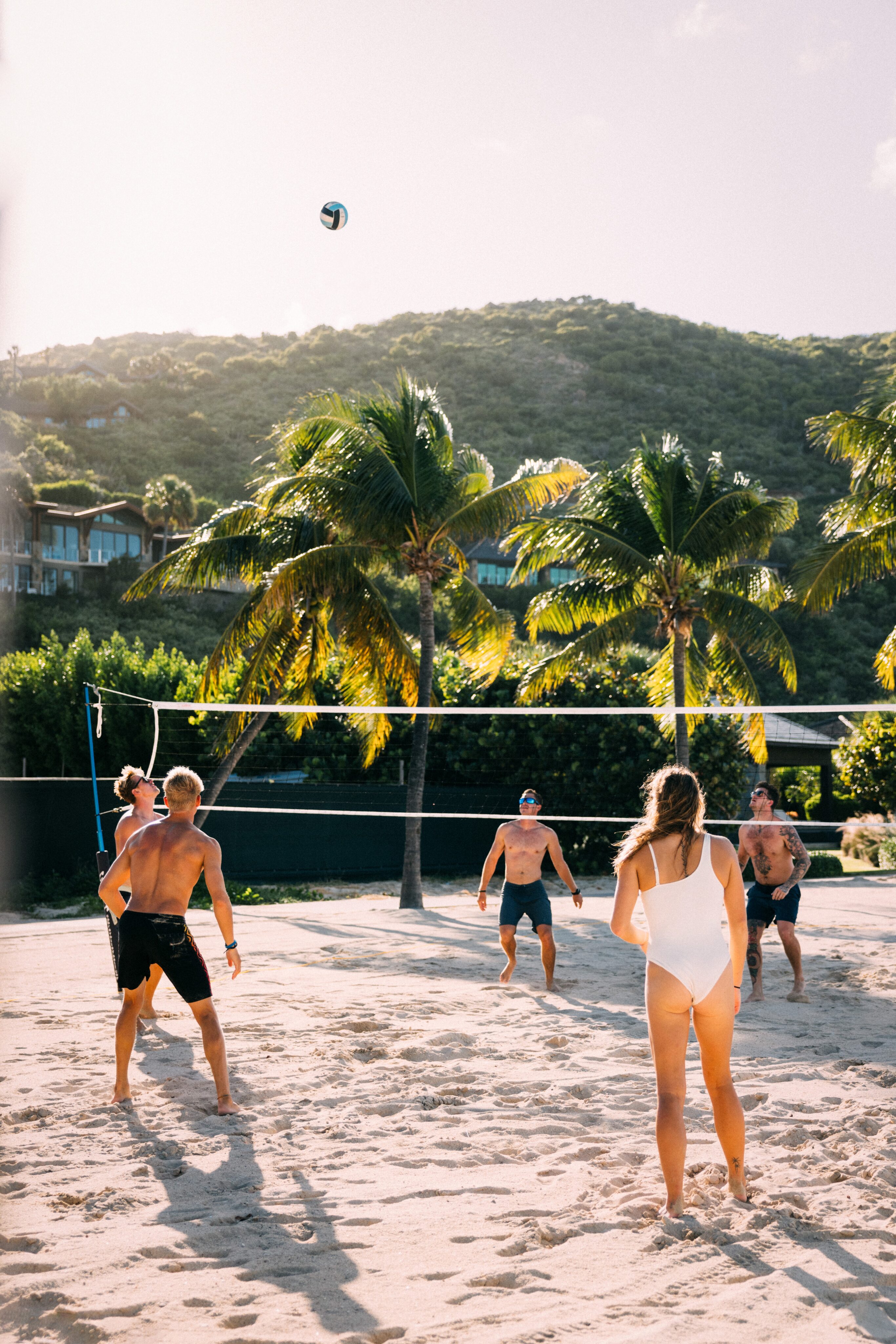 A group of people playing beach volleyball on a sandy court, surrounded by palm trees and green hills in the background. The sun is shining brightly, casting long shadows and creating a warm, vibrant atmosphere. One player in a white swimsuit is in the foreground, ready for action.