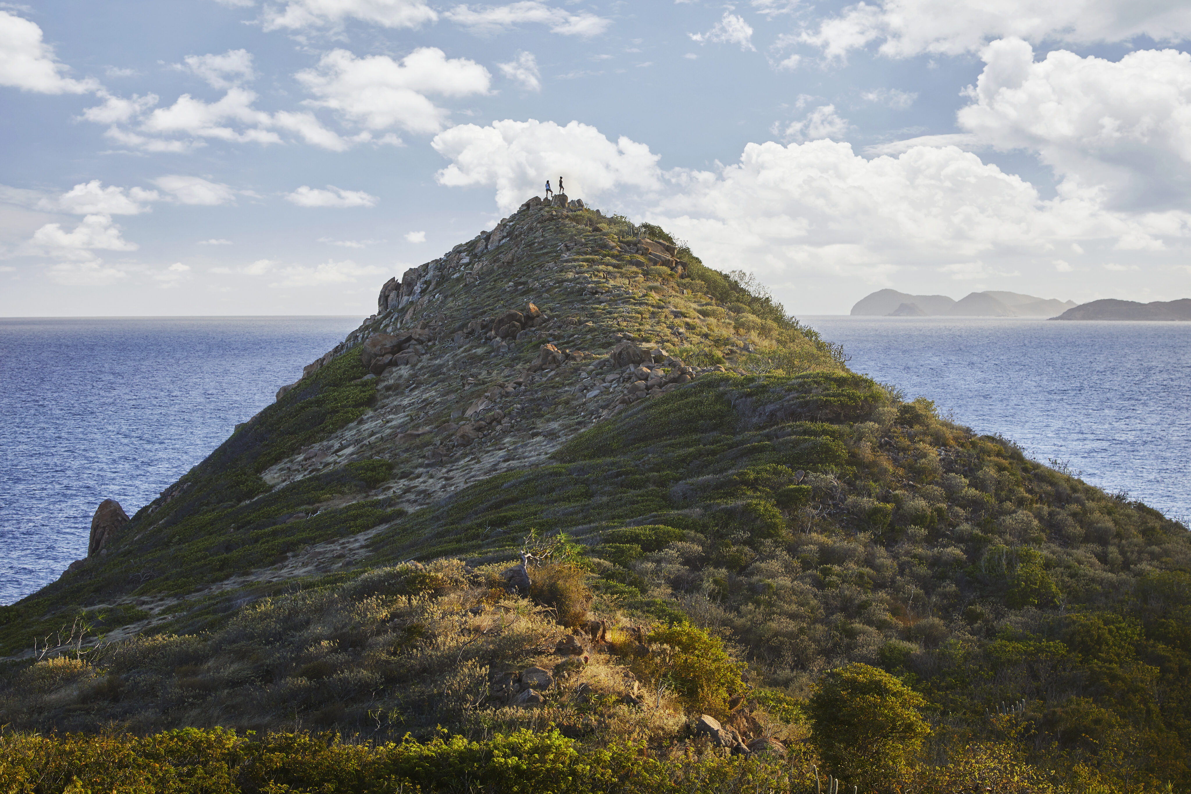 A group of people stands atop a rocky hill covered in sparse vegetation, enjoying outdoor activities while overlooking a vast ocean. The sky is partly cloudy, and distant islands are visible on the horizon. The hill slopes gently downward, with another hiker seen climbing halfway up.