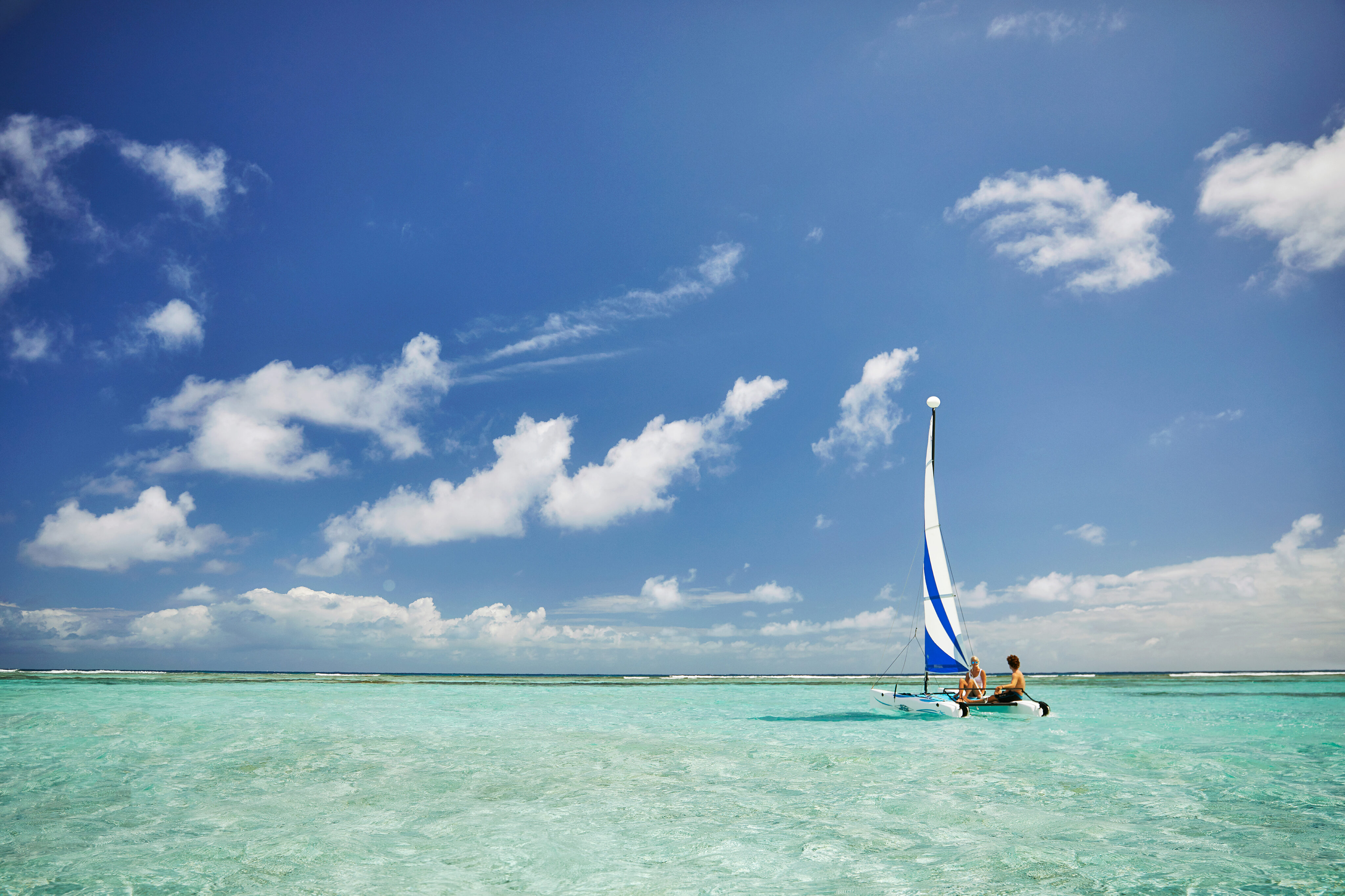 A small catamaran with a blue and white sail glides on clear, turquoise waters under a bright blue sky with scattered white clouds. Two people are visible on the boat, enjoying the serene and picturesque seascape.