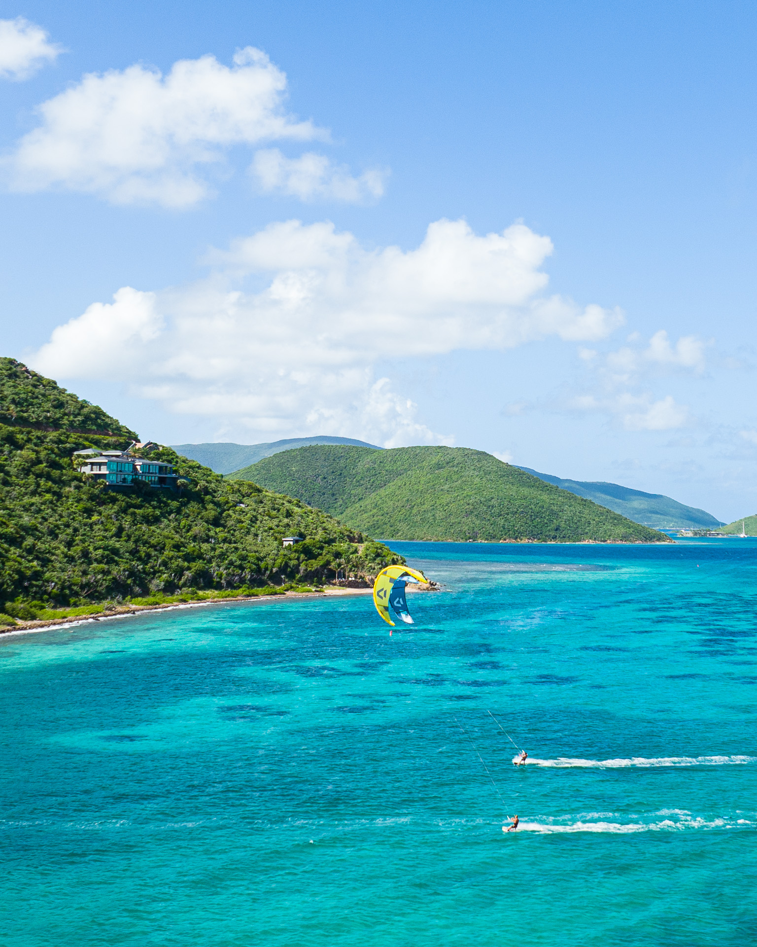 A scenic view of a vibrant, turquoise ocean alongside a lush, green hilly coast. A yellow parasail rises above the water, where enthusiasts enjoy kitesurfing, and two speedboats create wakes in the sea. Fluffy white clouds float in the bright blue sky.