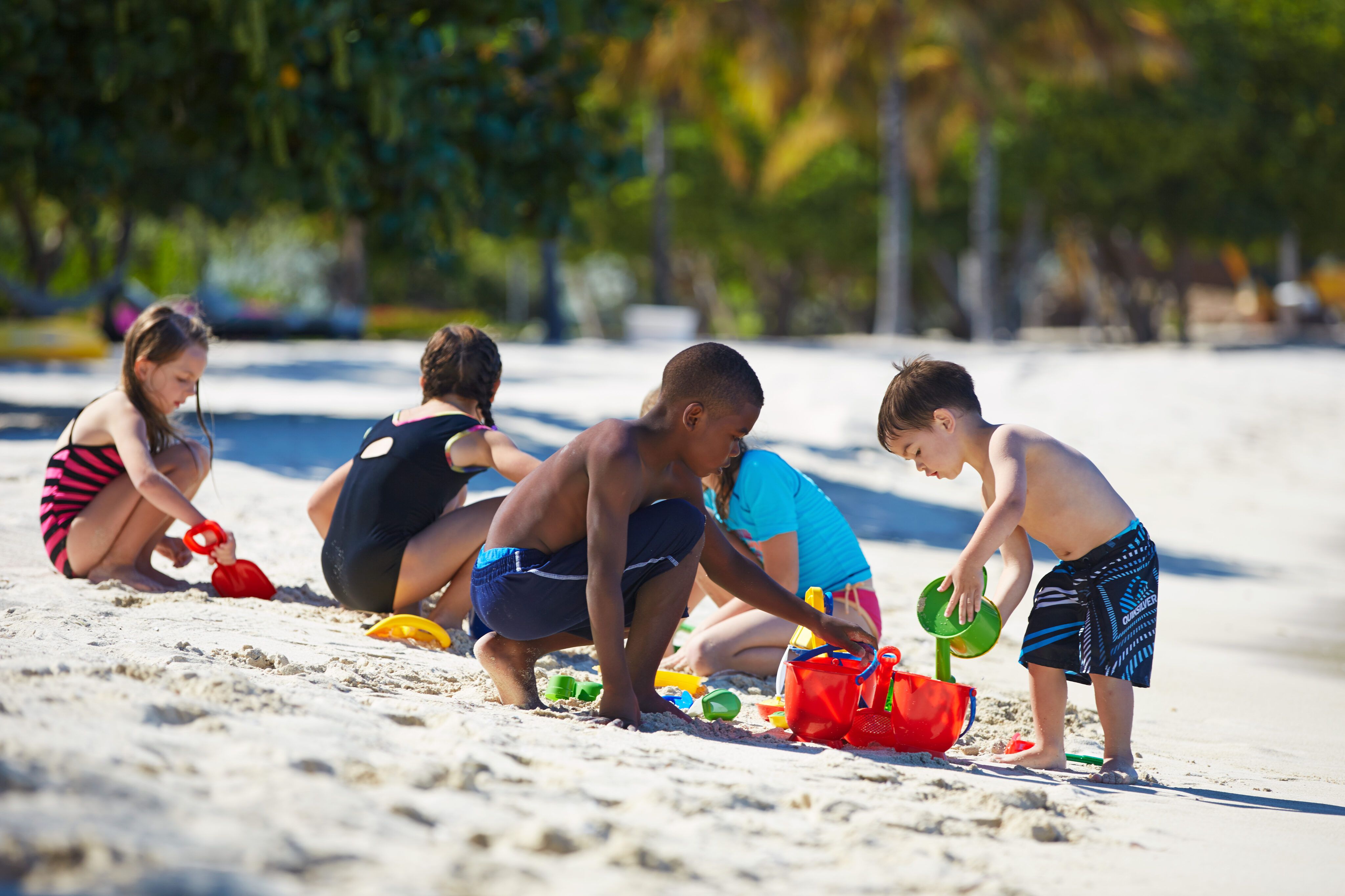 Children are playing together on a sandy beach. Two boys in swim trunks are building a sandcastle with colorful buckets and shovels, while two girls in swimsuits, supervised by a nanny agency's staff, engage in their own activities. Trees and foliage provide a lush backdrop.