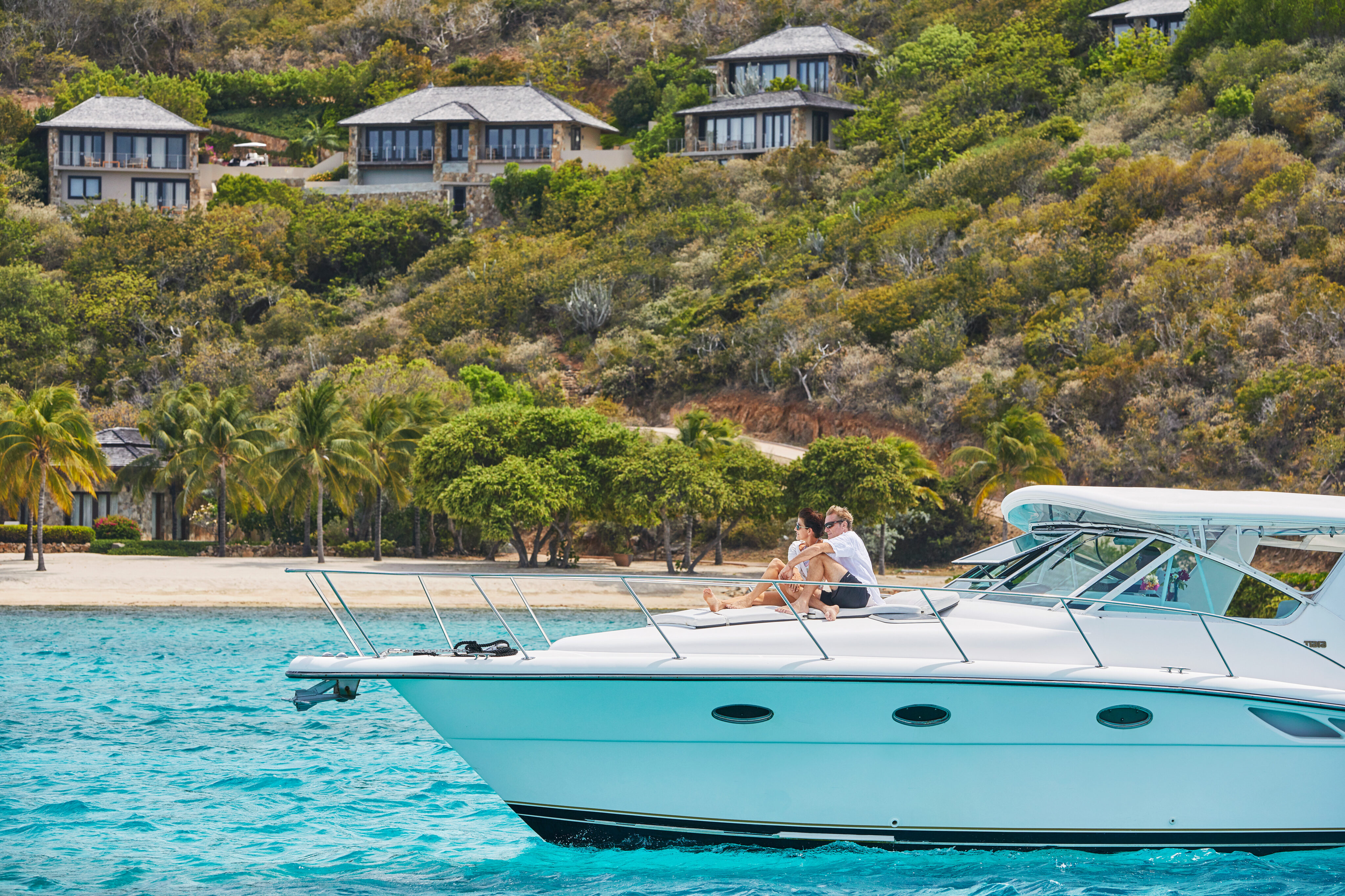 A couple sits on the bow of a luxury yacht charter in tropical turquoise waters, with a backdrop of lush greenery and beachfront villas. Palm trees line the sandy shore, and the scene depicts a serene and idyllic vacation setting.