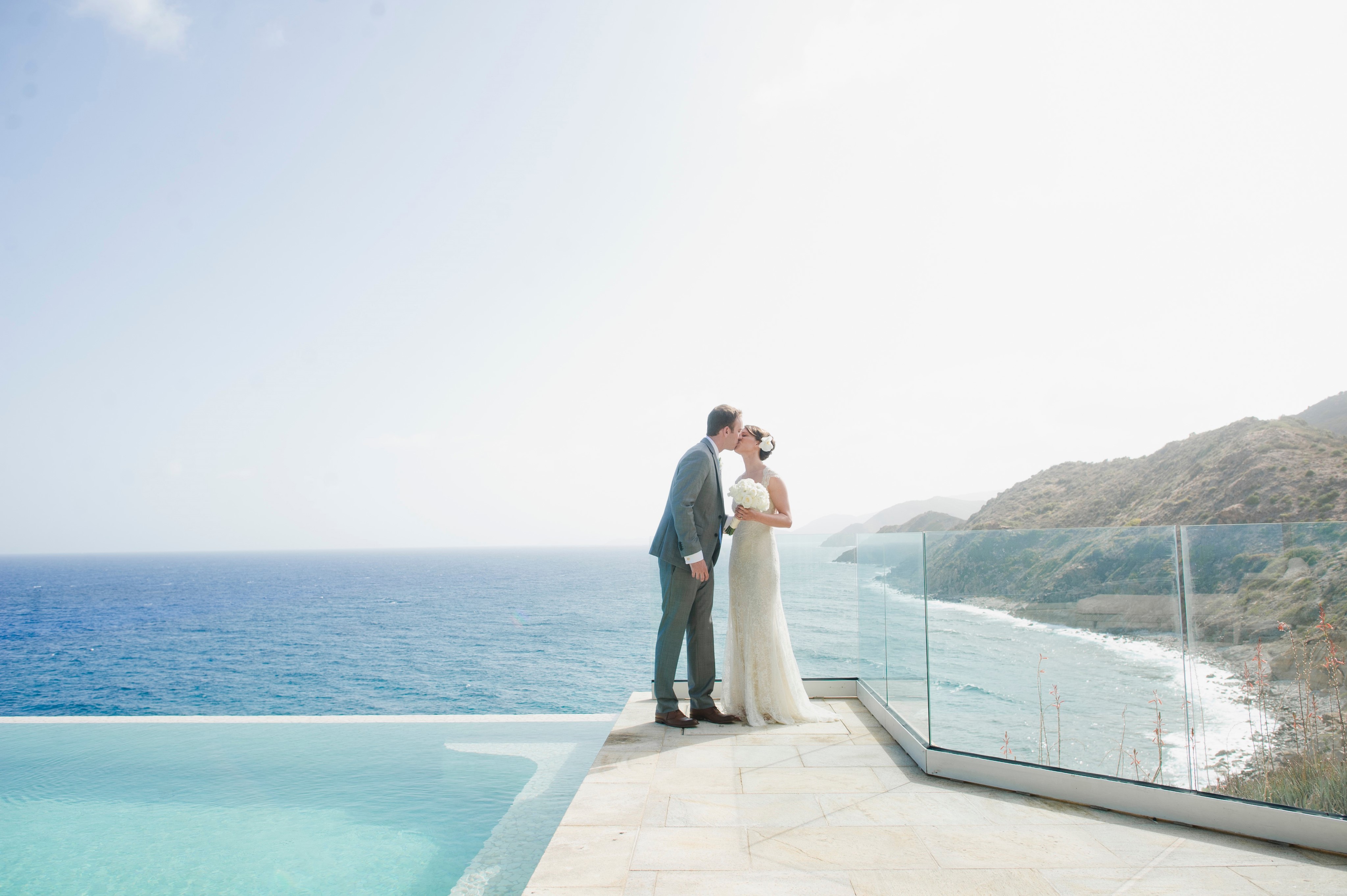 A couple, dressed in wedding attire, shares a kiss at the edge of an infinity pool that overlooks the ocean. The bride is in a white dress and holding a bouquet, while the groom wears a light suit. The background showcases a clear blue sea and rocky coastline, offering ideas for things to do in the BVI.