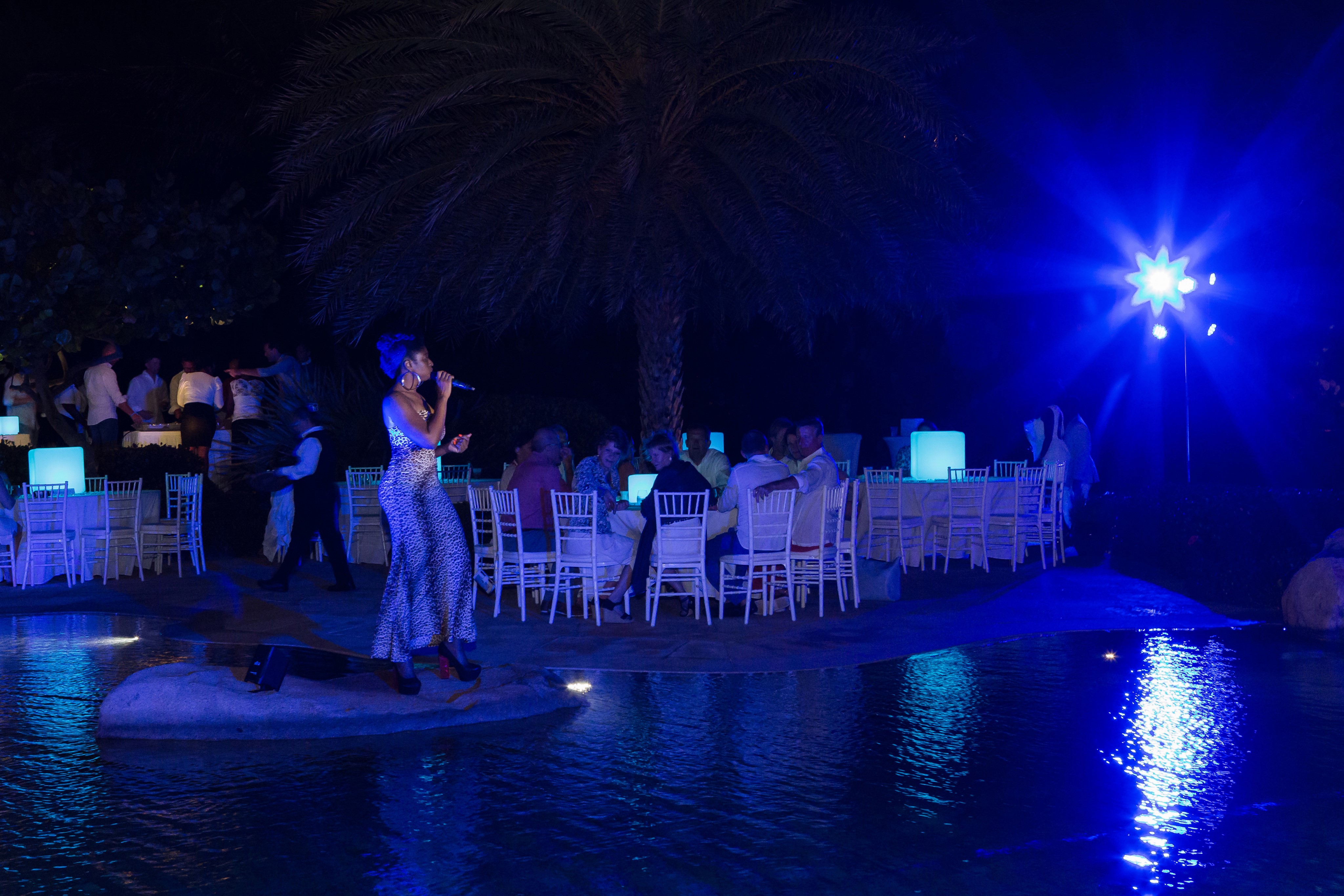 A performer with blue hair sings into a microphone on an outdoor stage surrounded by a pool. Guests seated at tables with glowing blue lights enjoy the evening. Palm trees are in the background, and a spotlight creates a vivid blue hue in the scene—a perfect highlight among BVI activities.