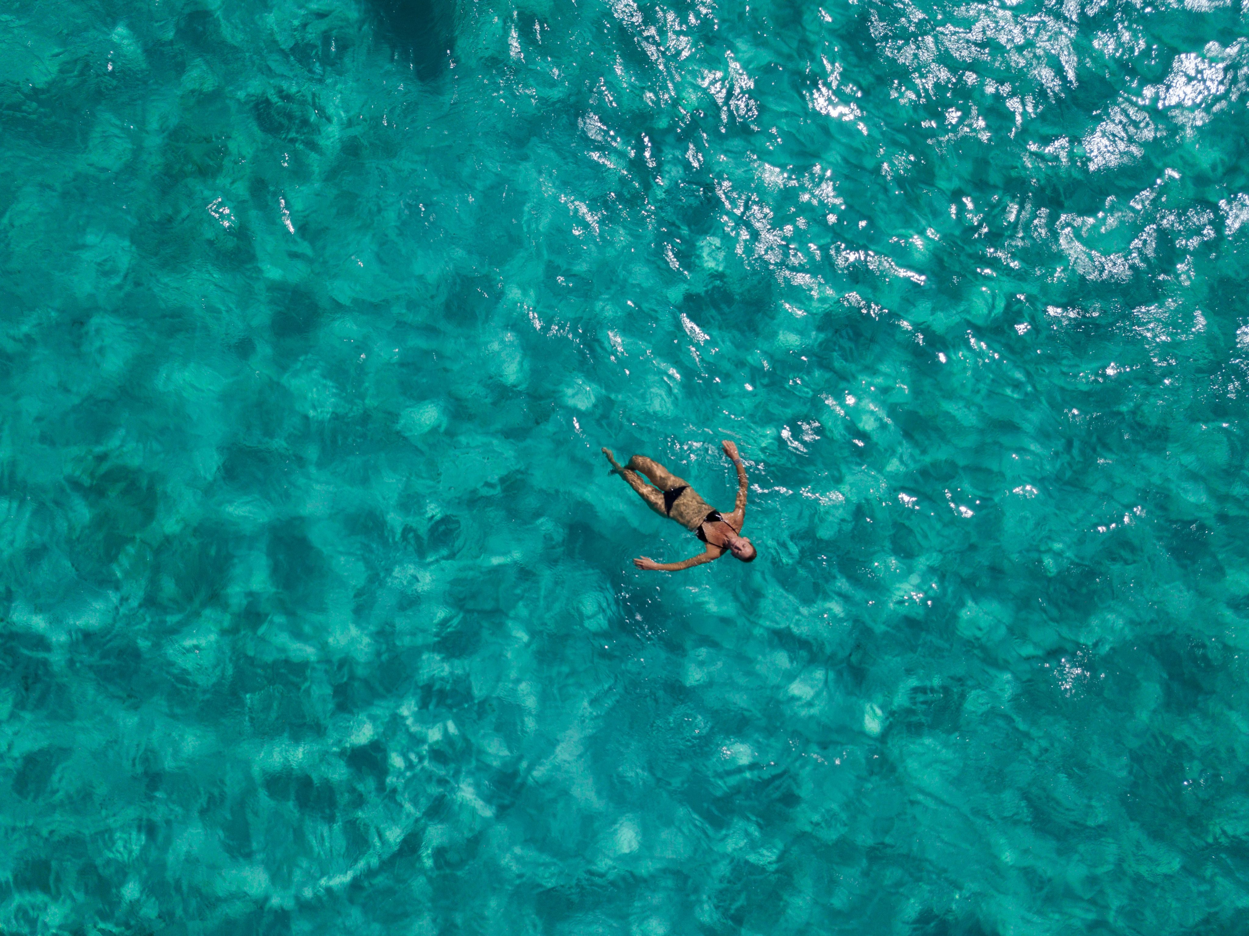 A person floats on their back in clear, turquoise ocean water. The water's surface glistens under the sunlight, creating a serene, tropical scene. The person is relaxed, arms outstretched, and appears to be enjoying the calmness of the sea—opting to stay in this moment of pure tranquility.