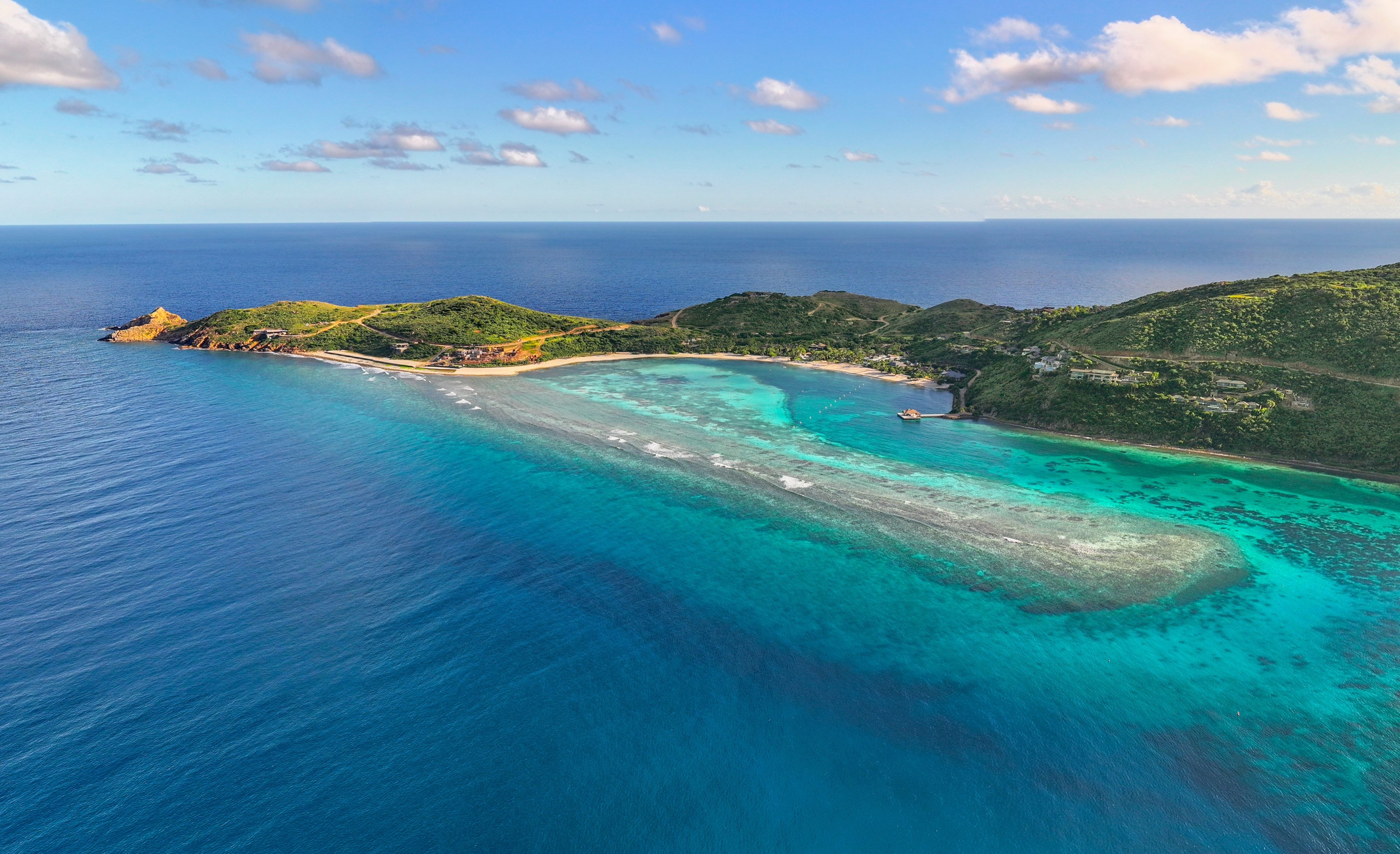 Aerial view of Oil Nut Bay, a tropical island surrounded by clear turquoise and deep blue waters. The island features lush green hills and sandy beaches, with some rocky areas. A few scattered structures are visible near the shoreline under a partly cloudy sky.