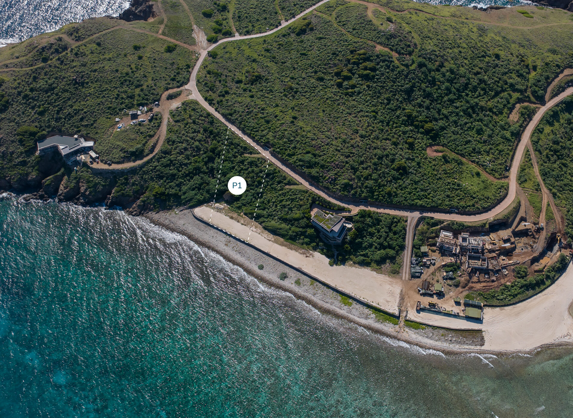 Aerial view of the Pirogue Beach at Saba Rock Resort showing lush green hills, winding roads, and several homesites scattered on the landscape. The clear blue ocean borders the beach, with prominent markers labeled "P1" and "P2" on the map close to the peninsula shoreline.