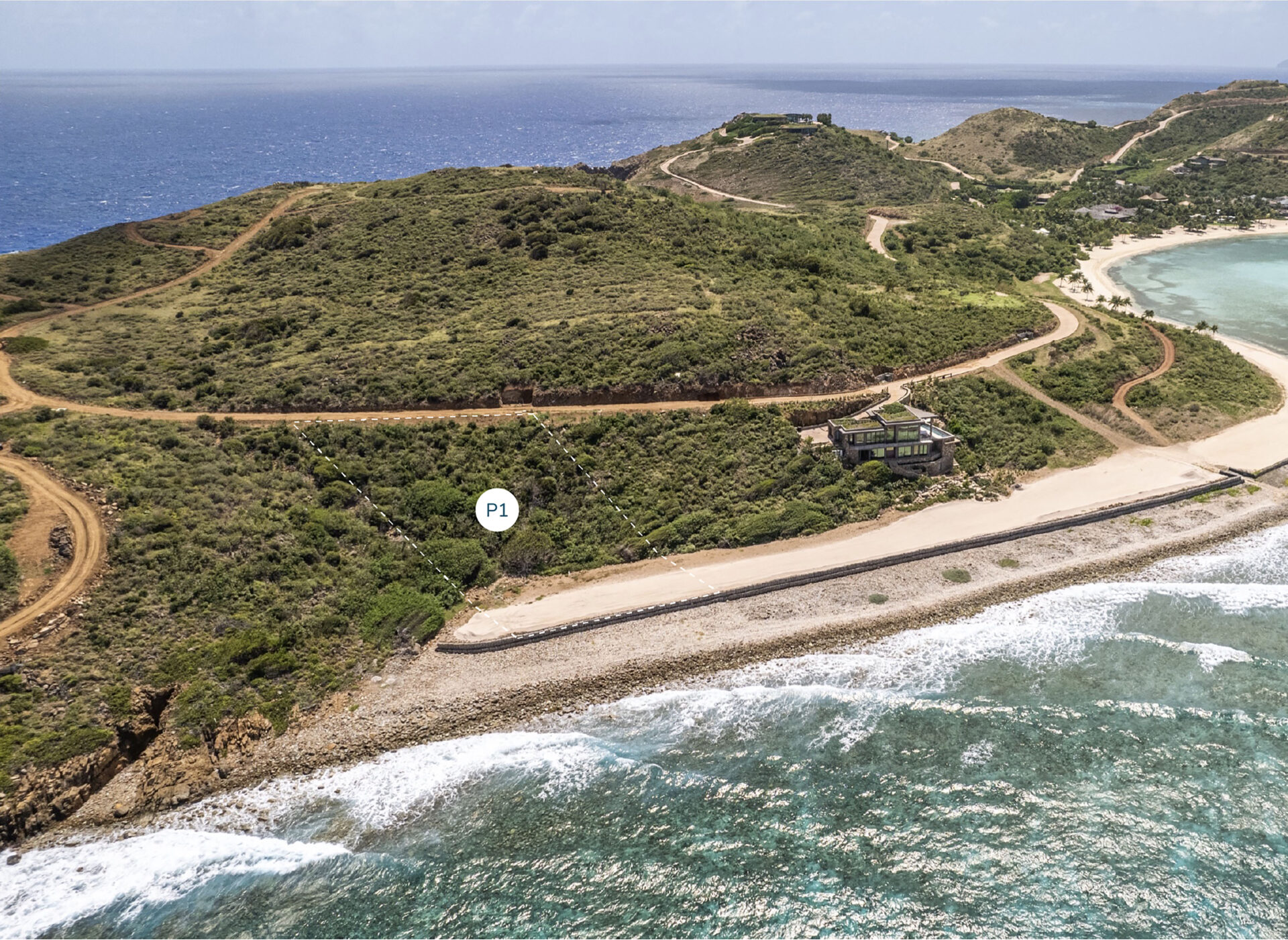 Aerial view of a coastal landscape featuring a winding road atop green hills, a sandy beach with gentle waves, and blue ocean in the background. An area marked "P1" is visible near the beach alongside where Peninsula Homesite is noted further inland. Sparse vegetation dots the hills, and a single building is visible along the road.