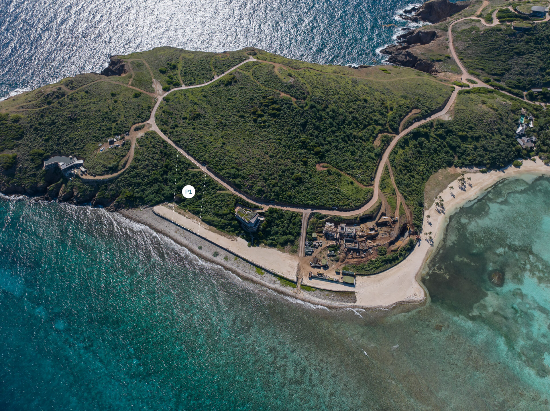 Aerial view of a coastal landscape with winding paths on a grassy hill leading down to a beach. Ruins of old structures are visible near the shoreline, marking the historic Peninsula Homesite. The surrounding water is clear with shades of blue and turquoise, and waves lap gently against the sandy shore.