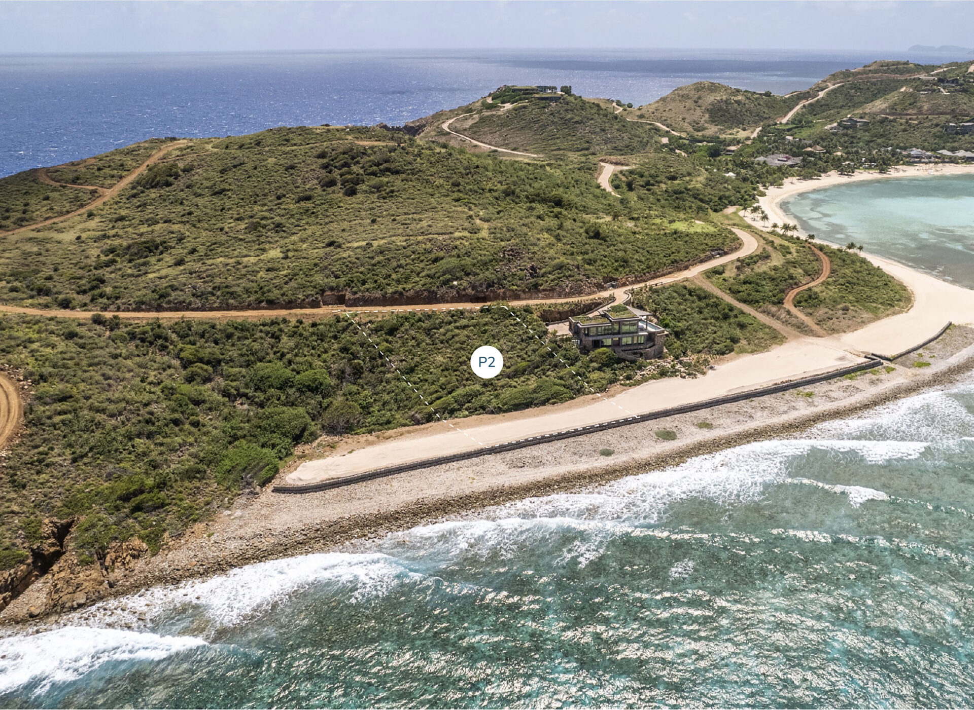 Aerial view of a coastal landscape with a hilly terrain. The photo shows a rocky shoreline bordered by lush green hills. A winding road traverses the hills, leading to Peninsula Homesite P2, centrally located in the image and labeled clearly as "P2.