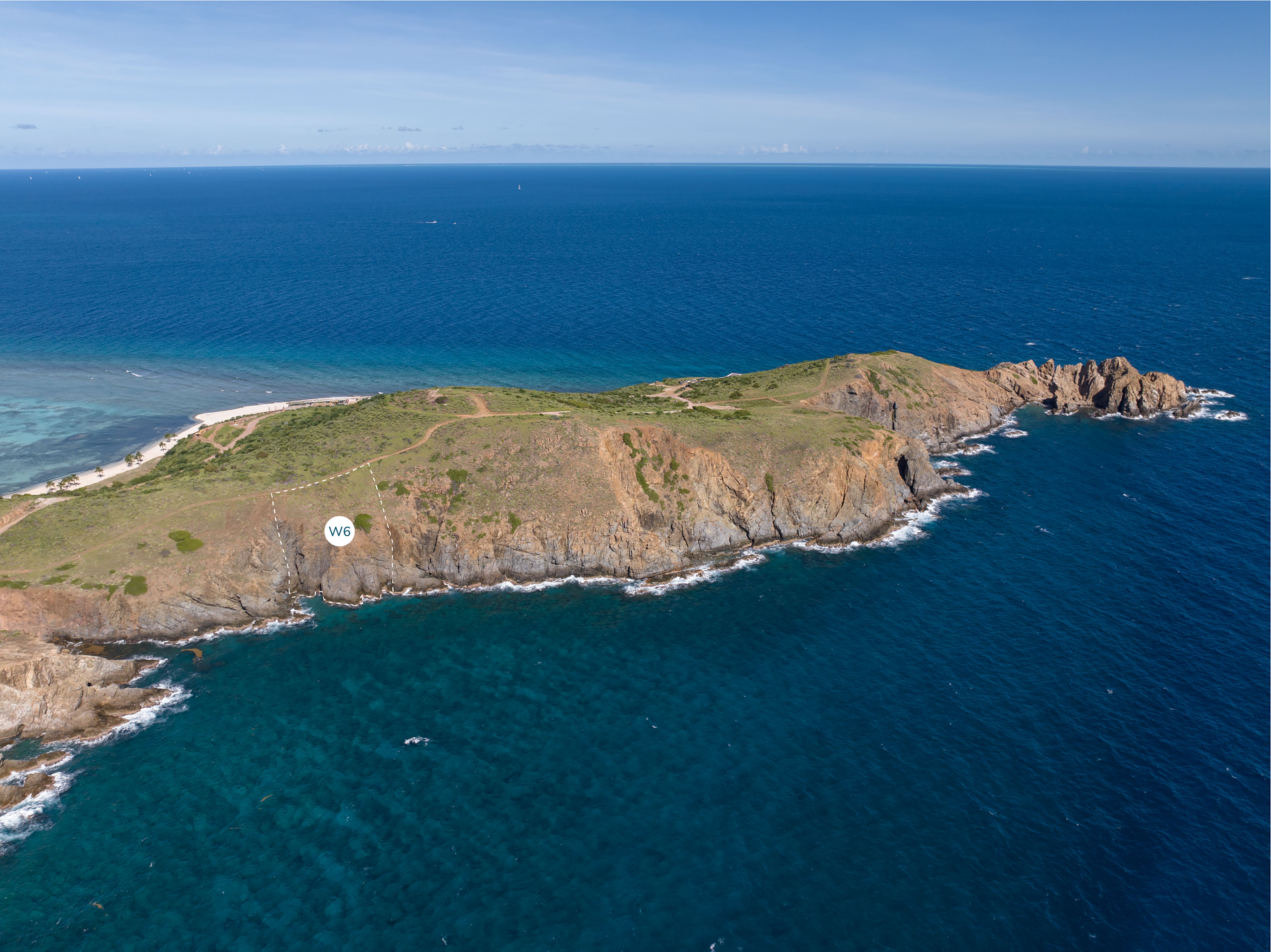 Aerial view of a rocky coastline with green vegetation atop cliffs meeting the blue ocean. There is a marker labeled "Homesite W6" placed on one of the cliffs near the shoreline. The horizon shows a clear sky blending into the expansive ocean, perfectly capturing this Wildside retreat.