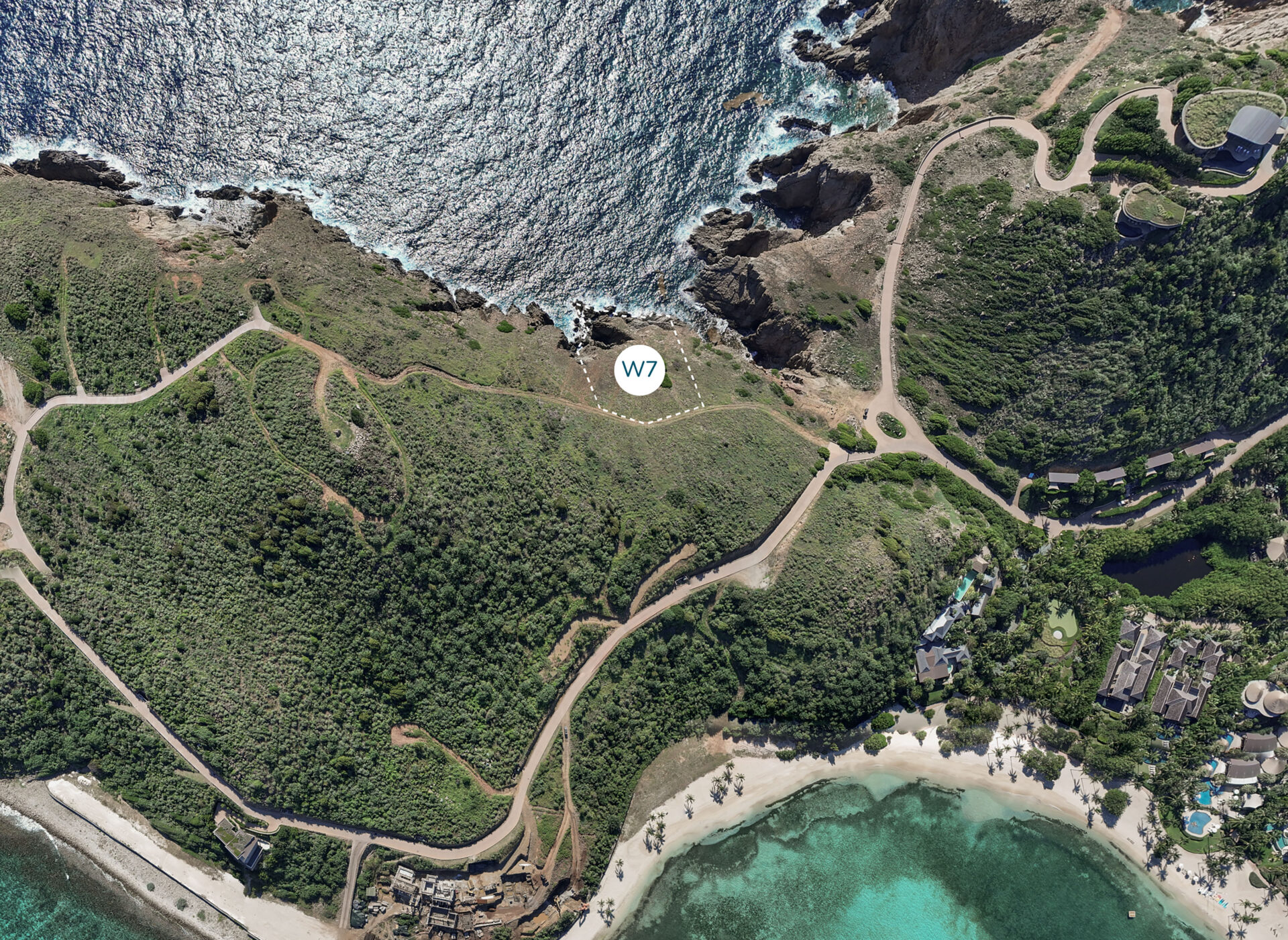 An aerial view of a coastal area featuring a lush green landscape with winding roads, rocky cliffs, and a turquoise bay. The area is marked with a white circle labeled "Homesite W7." The coastline boasts clear water, sandy beaches, and scattered buildings near Wildside Homesite.