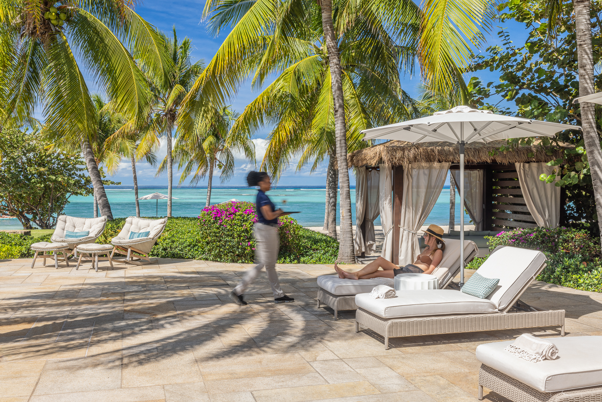 A woman relaxes on a lounge chair under a white umbrella by the beach, surrounded by tropical palm trees. Another person walks by, holding a tray. The clear blue ocean and sky are in the background, along with a thatched-roof cabana and additional lounge chairs.