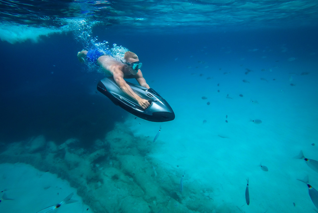 A person wearing swim trunks and goggles rides an underwater scooter, enjoying an exhilarating seabobbing experience through a clear, blue ocean, surrounded by fish and rocks on the sea floor.