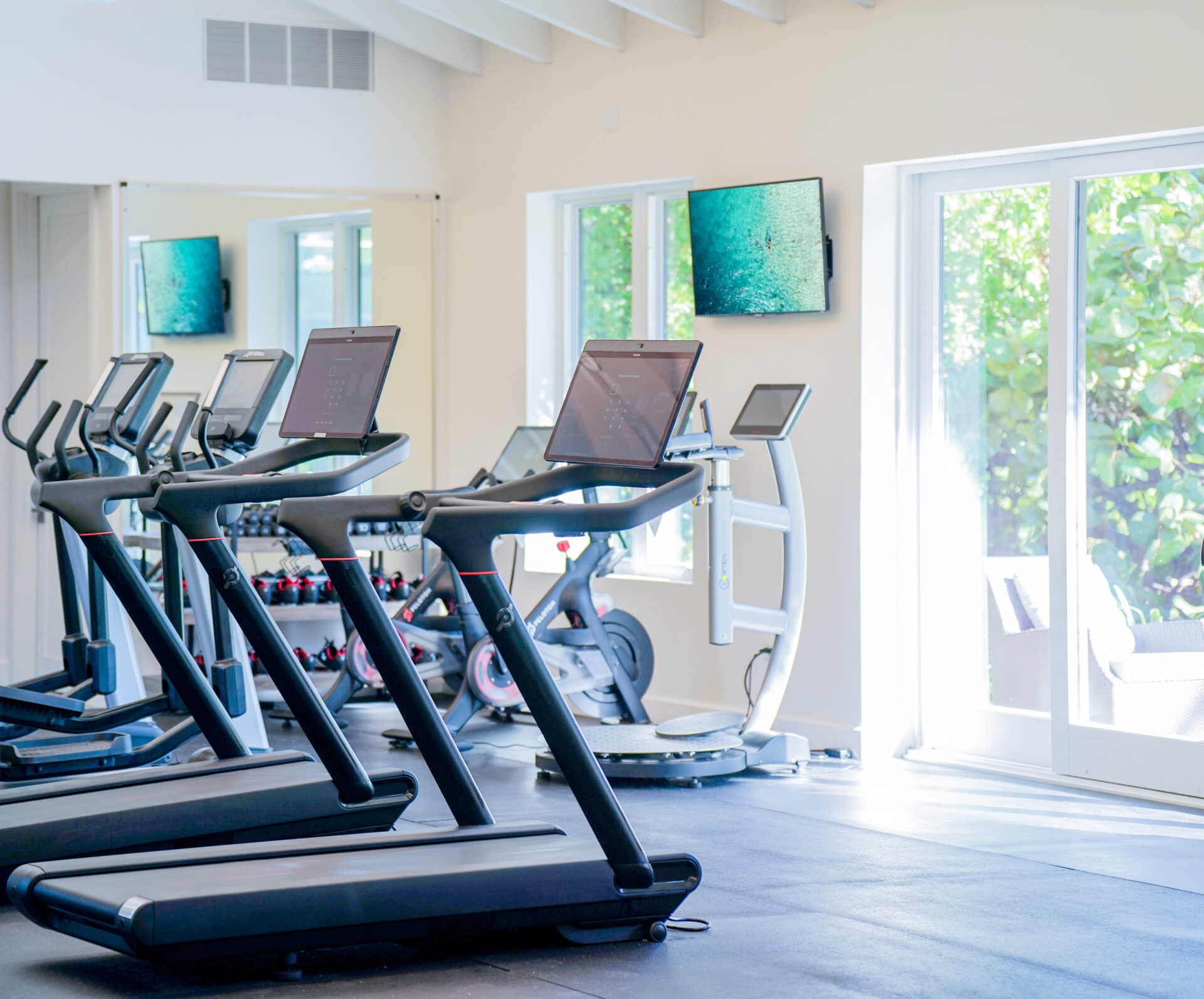 A well-lit health studio with several treadmills facing a wall-mounted TV and mirrors. There are large windows on the right letting in natural light, promoting wellness. A stationary bike and other equipment are in the background.