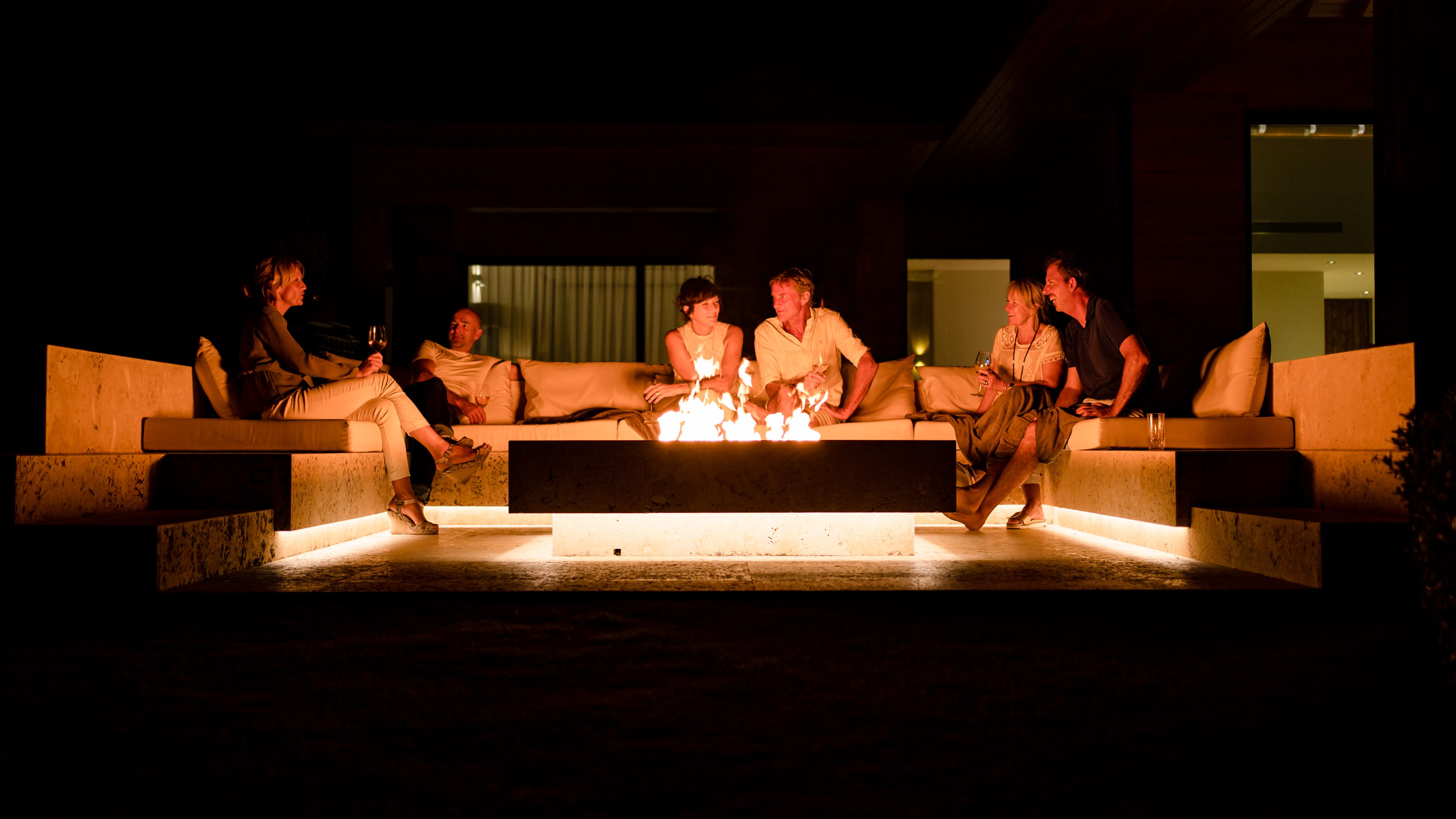 A group of people sits on beige couches around a rectangular fire pit at night. The outdoor seating area is well-lit with soft, warm lighting emanating from the fire pit and underneath the seating. The scene appears relaxed and social.