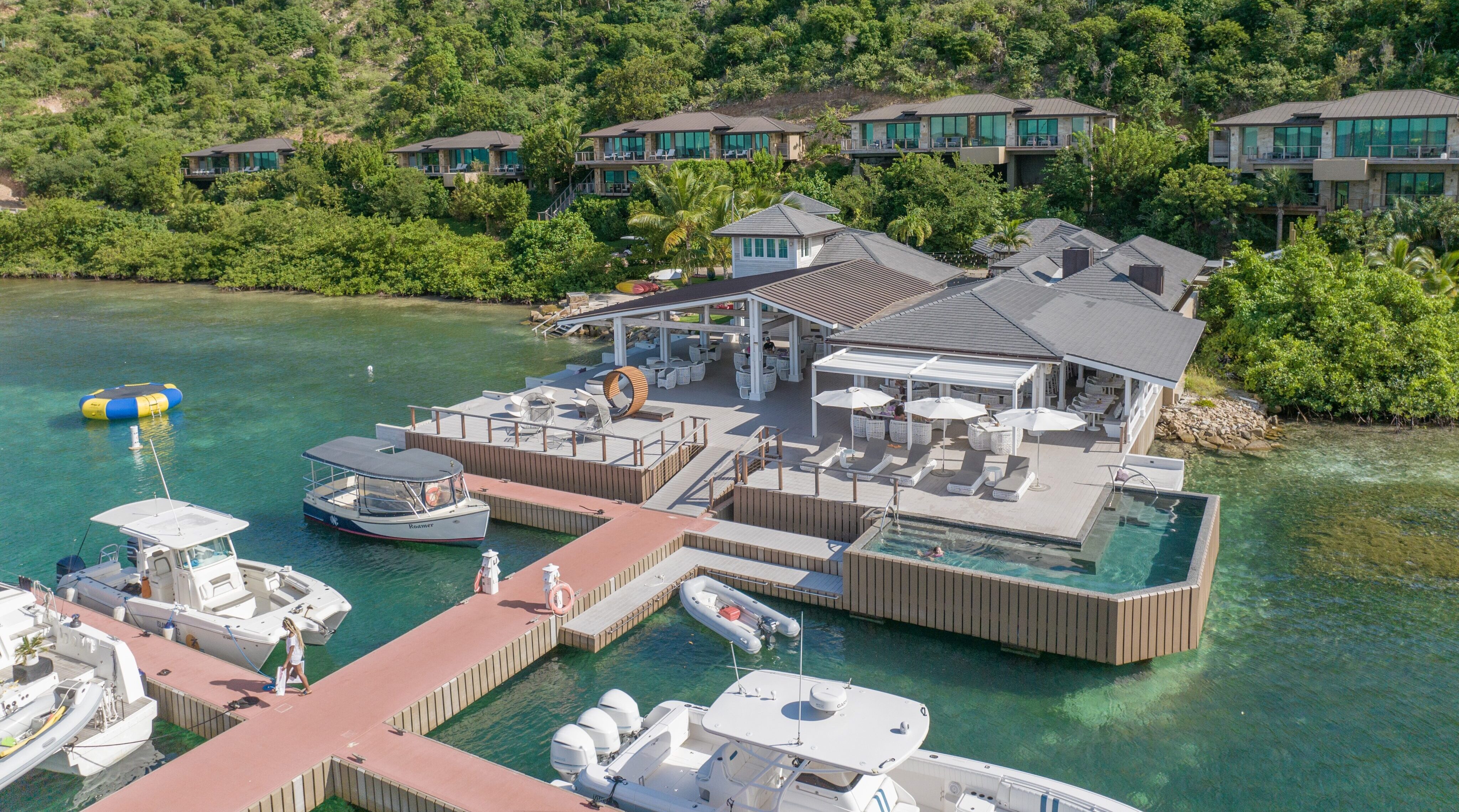 A scenic view of a marina village in the British Virgin Islands, with several boats moored in clear blue water. Nearby, a modern waterfront building with outdoor lounging areas sits against a backdrop of lush green hills and several elevated cabins nestled in the foliage.