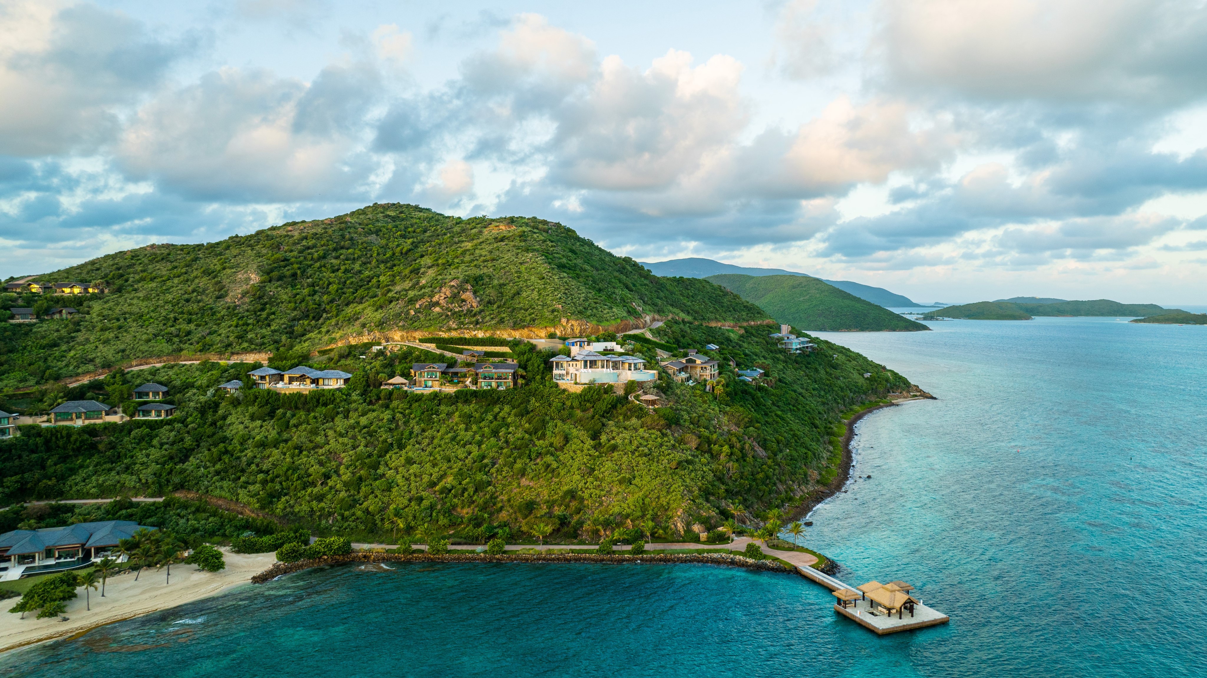 A scenic drone view of lush, green hilly coastline dotted with luxury real estate atop the hills. The turquoise ocean stretches out, meeting a wooden dock extending into the water. Clouds scatter across the blue sky, capturing the serene beauty of Oil Nut Bay in the British Virgin Islands.