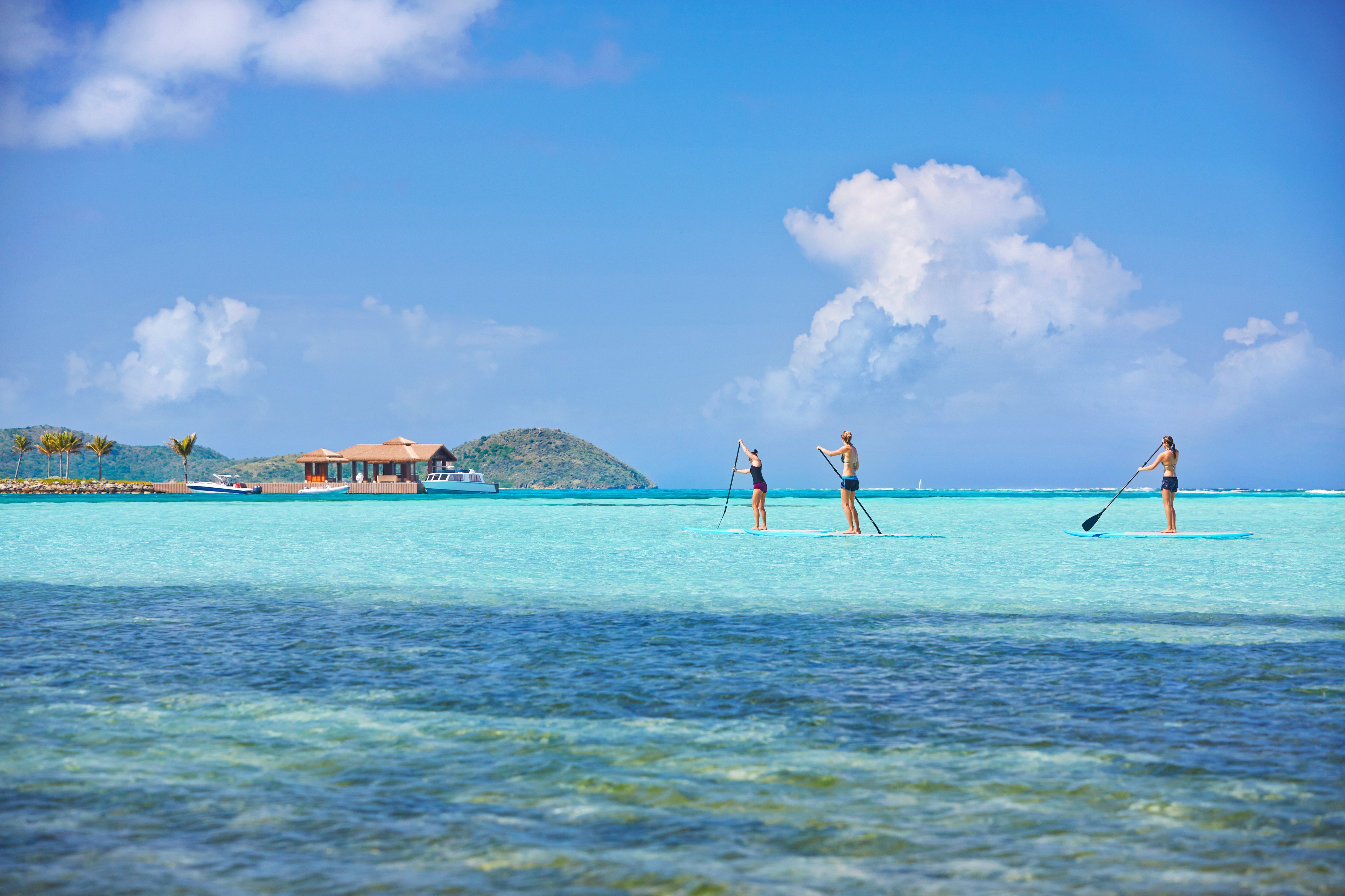 Three people are stand-up paddleboarding on clear turquoise water under a bright blue sky with a few clouds. In the background, there is a small house on a hill beside the water, part of the exclusive luxury real estate available at Oil Nut Bay in the British Virgin Islands, surrounded by lush green hills.