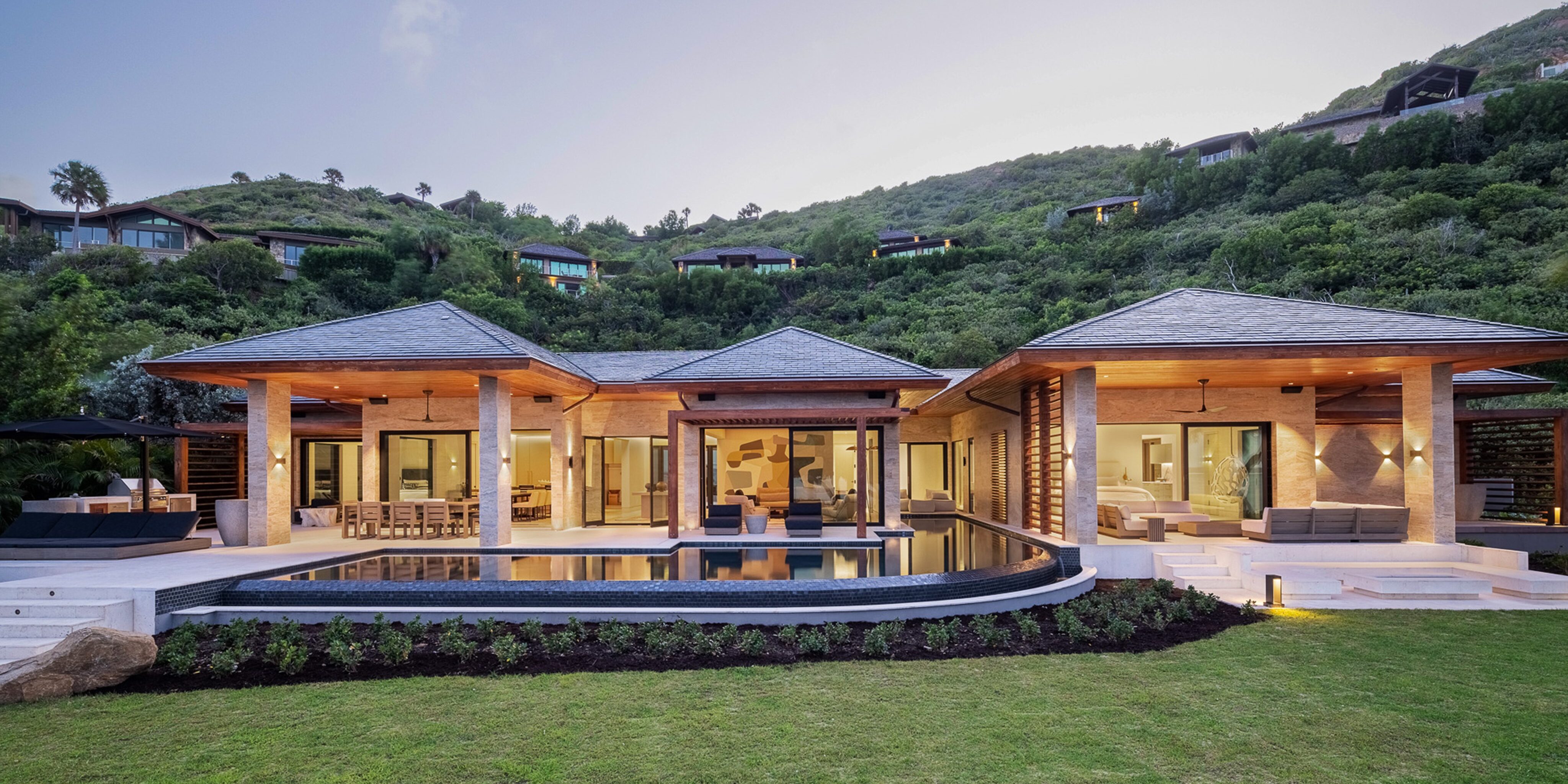 A luxurious modern house for sale in the British Virgin Islands' exclusive Oil Nut Bay, with three sections interconnected by outdoor walkways, features expansive glass windows, a large infinity pool, and a spacious lawn. It's set against a lush hillside with additional homes visible in the background.