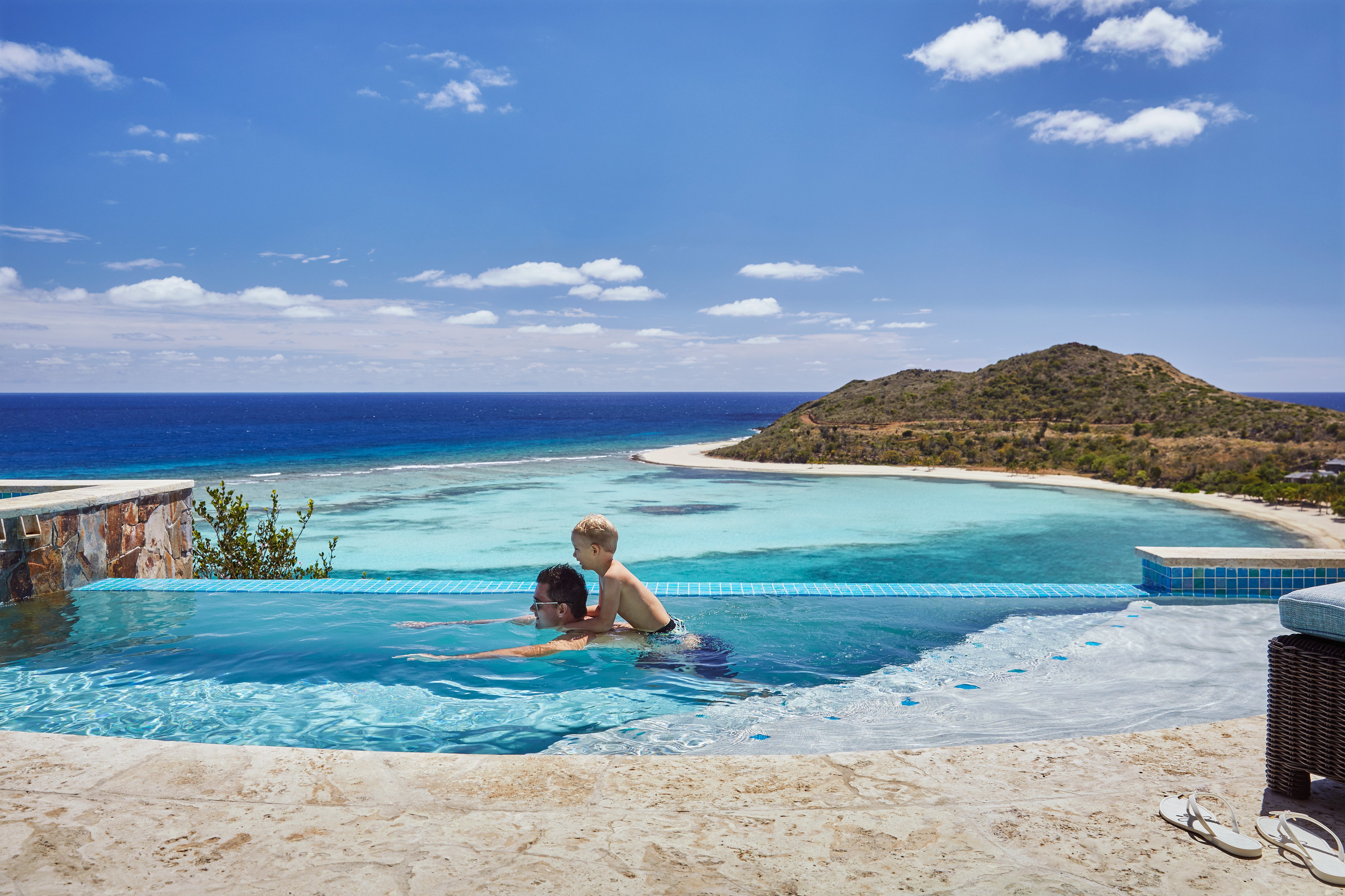 A man and a young boy play in an infinity pool overlooking a pristine turquoise ocean at Oil Nut Bay, a premier destination offering luxury real estate in the British Virgin Islands. The pool edge blends seamlessly into the horizon, with a sandy beach and a small, green hillside in the distance. The sky is bright blue with scattered clouds.