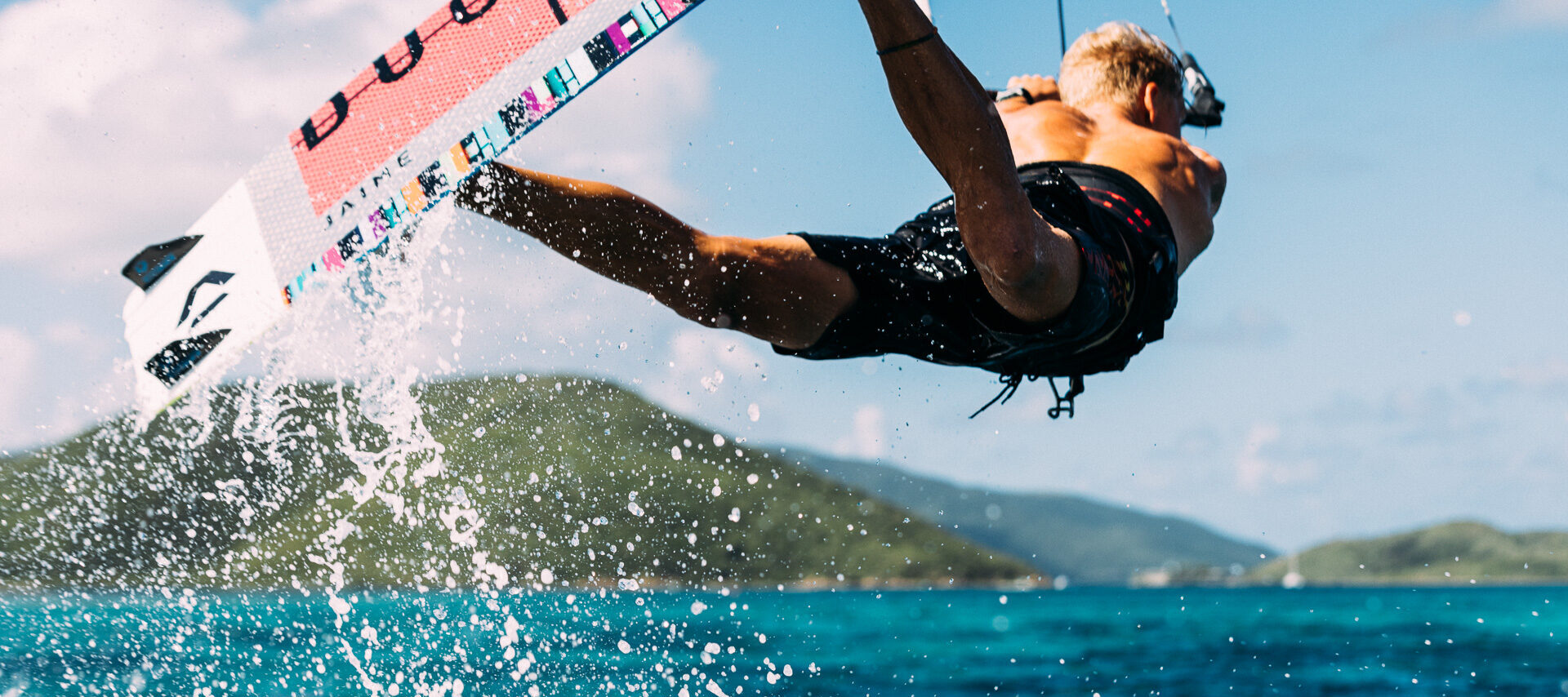 A person is kiteboarding over the ocean near Marina Village in the British Virgin Islands, with mountains in the background. The individual is airborne, holding onto the kite, and water splashes around them under a clear, blue sky. They are wearing black shorts, and the kiteboard features colorful graphics.