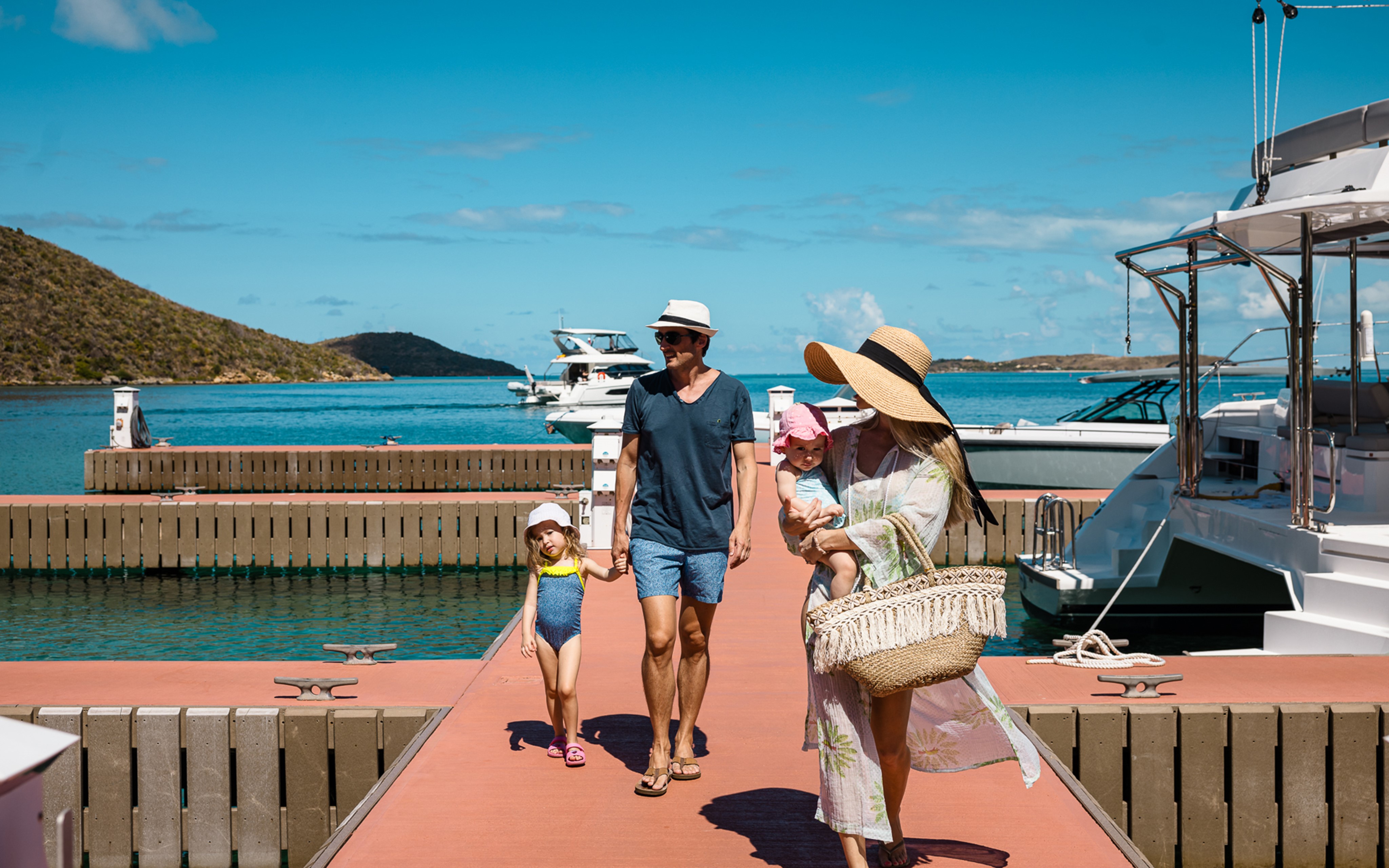 A family of four walks on a dock beside yachts and clear blue water in Marina Village in the British Virgin Islands. The parents hold hands with their two young children, who are wearing hats, while mom carries a woven basket. The sky is bright blue with scattered clouds, and a hill is visible in the background.