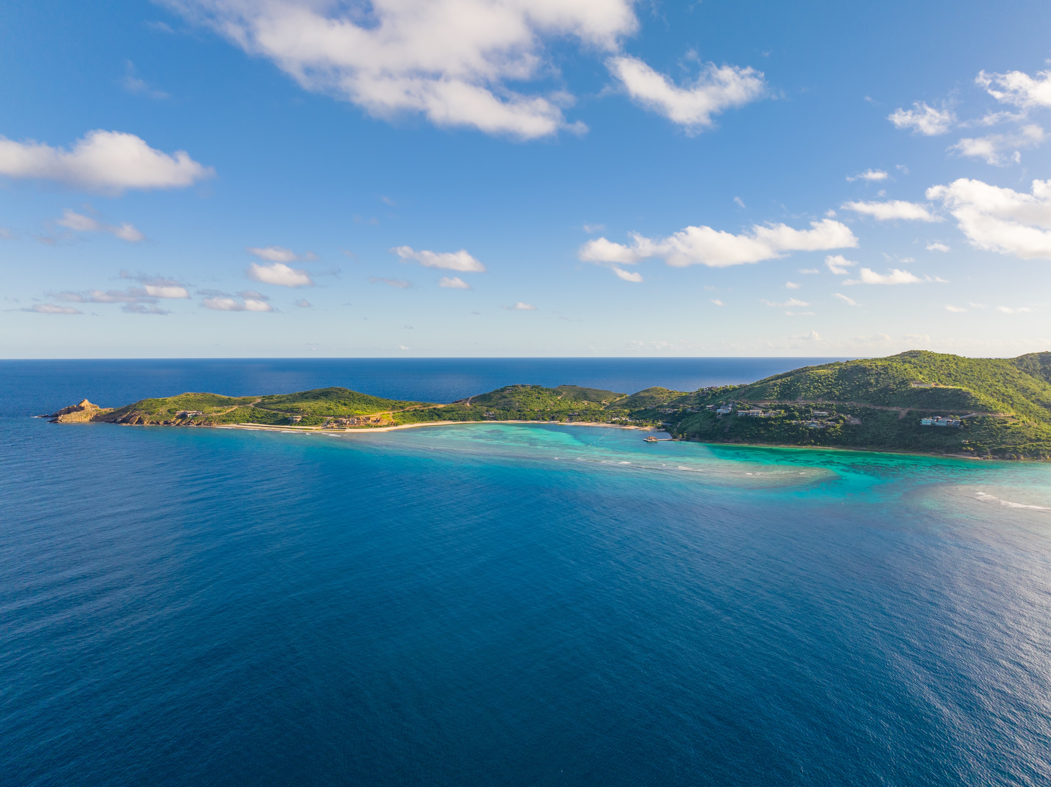 Aerial view of a serene coastline at Oil Nut Bay, with bright blue water, lush green hills, and scattered clouds in a clear sky. The luxury real estate boasts a mix of vegetation and coastline, surrounded by the vast expanse of the ocean. The horizon is visible in the distance.