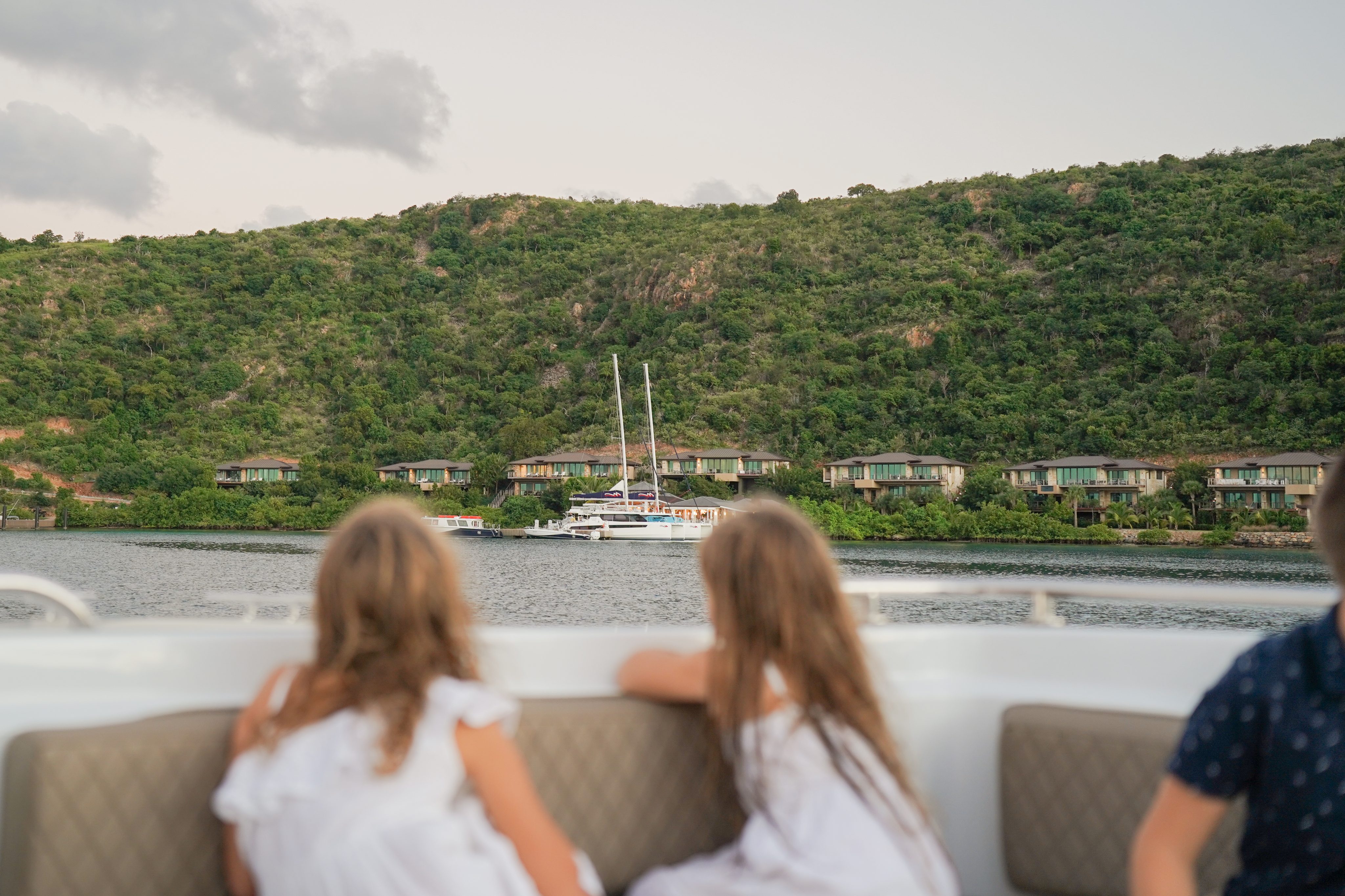 Two girls in white dresses sit on a boat, gazing towards a lush green hillside dotted with houses and a large sailboat anchored near the shore in the Marina Village in the British Virgin Islands. The water is calm, and the sky is overcast with a few clouds.