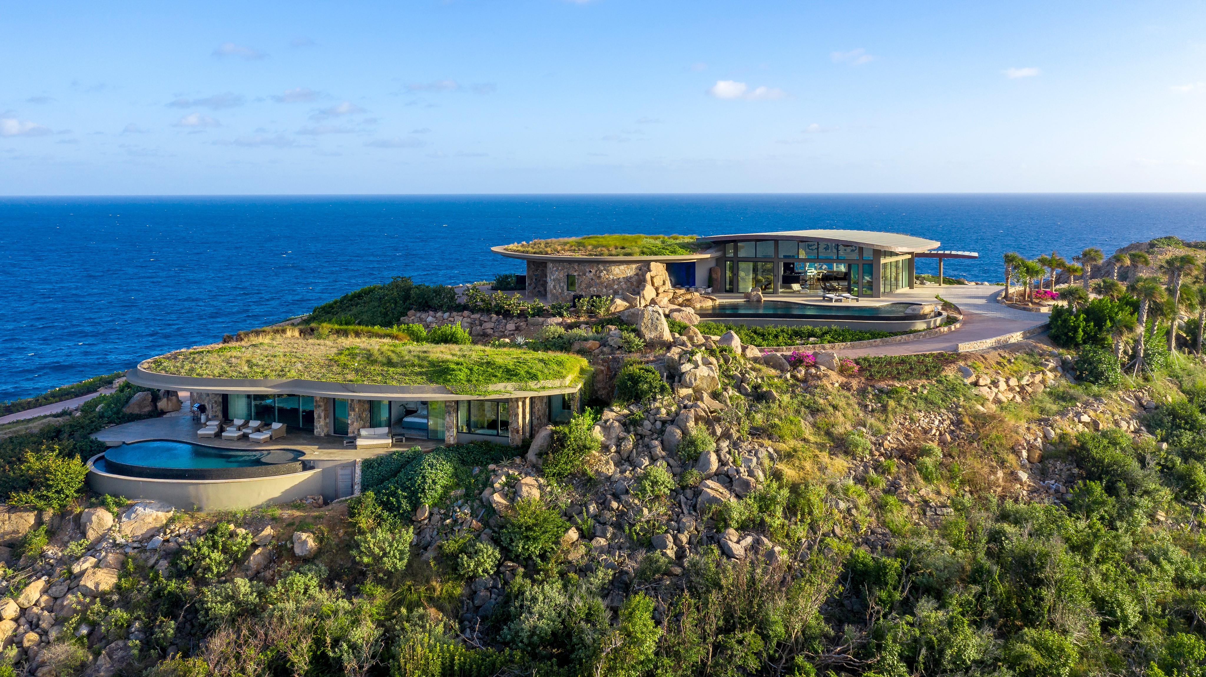 Aerial view of two modern, eco-friendly houses with green roofs on a rocky coastline overlooking a vast blue ocean. About the landscape: lush vegetation surrounds, a pool at the nearest house shimmers under the mostly clear sky with a few scattered clouds.