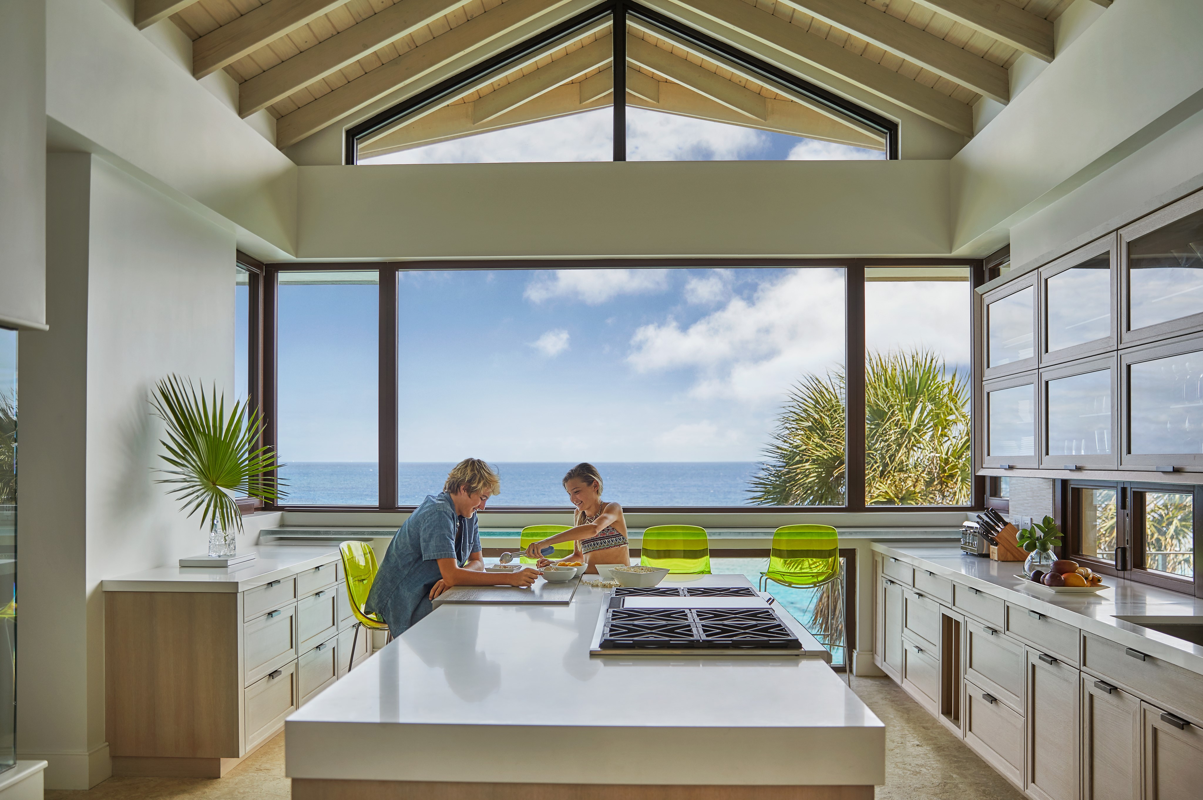 A spacious, modern kitchen with large windows overlooking the ocean. Two people sit at a white island counter; one is writing, and the other is observing. Bright green chairs add a pop of color, and a palm frond decorates the counter. About them, the sky is clear and sunny outside.