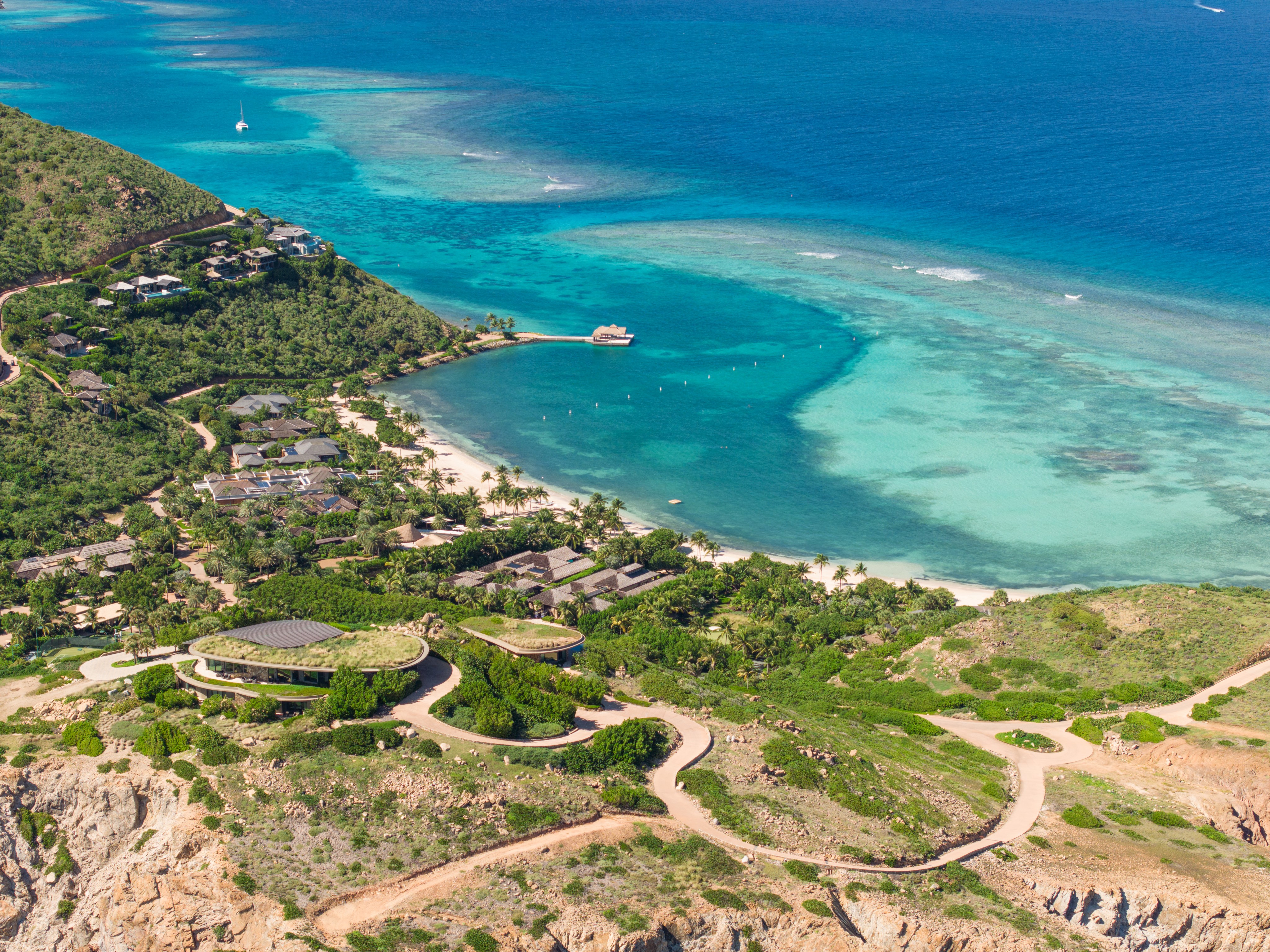 An aerial view reveals a coastal resort area featuring green hills, modern architecture, lush vegetation, and clear turquoise waters with sandy beaches. Scattered throughout the landscape are paths and buildings, with boats visible in the water. It's all about relaxation and natural beauty.