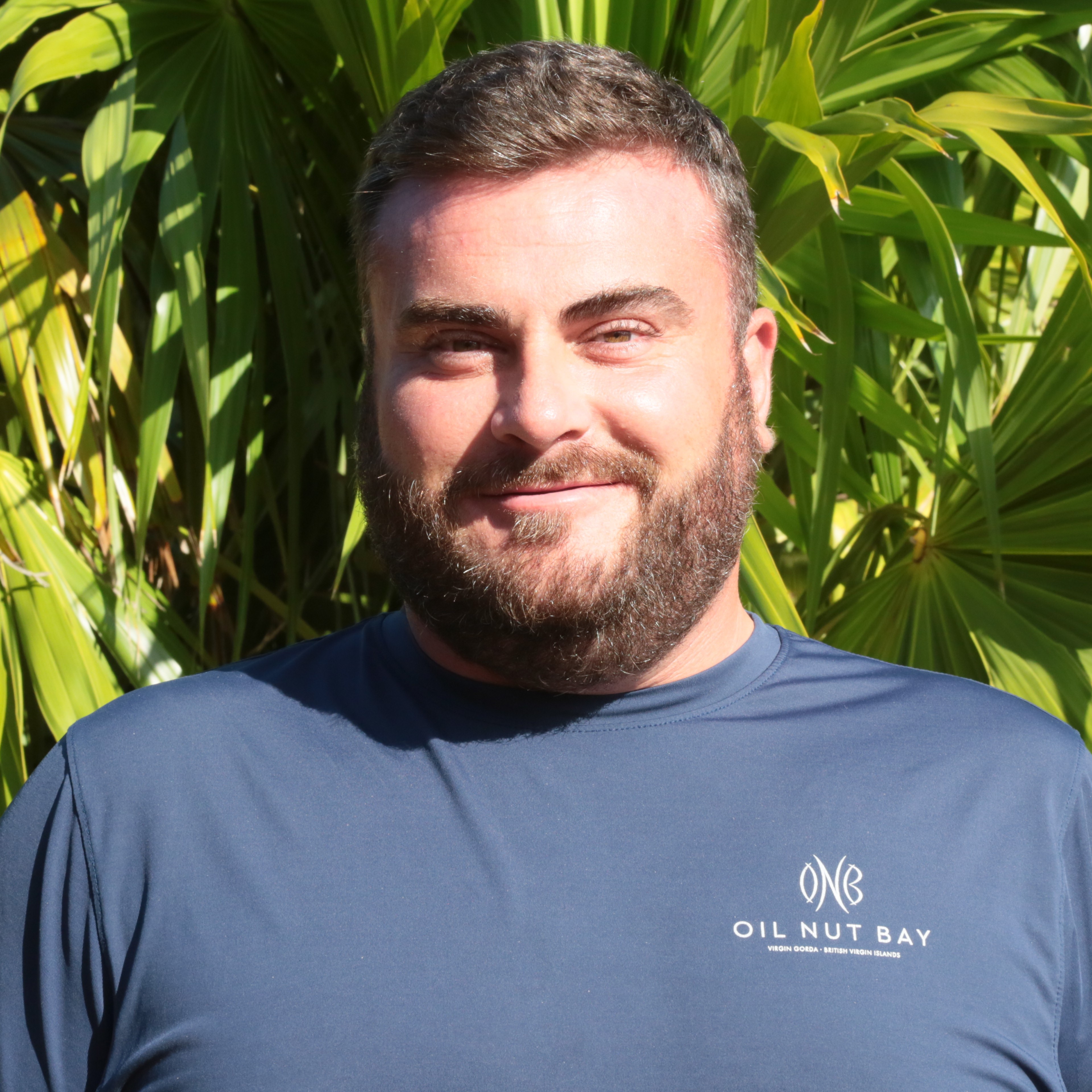 A man with a beard, Brendan O'Reilly, stands in front of a backdrop of green palm leaves. He is wearing a blue shirt with the text "OIL NUT BAY" and a logo on it. With a faint smile, he looks directly at the camera.