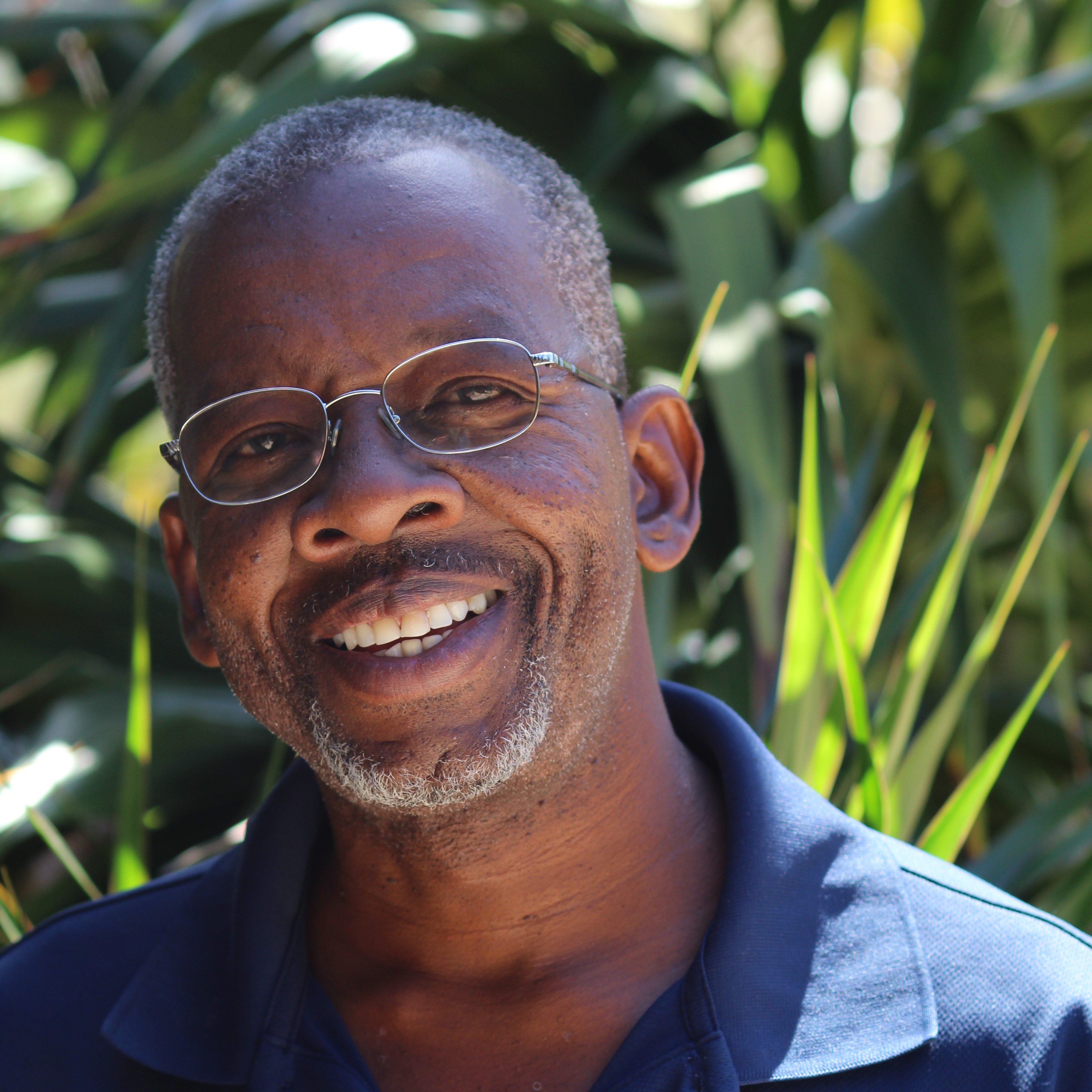 A smiling man with glasses and a short beard, wearing a blue polo shirt, is standing outdoors with greenery in the background. Clyde Lettsome's serene presence radiates through the natural setting.