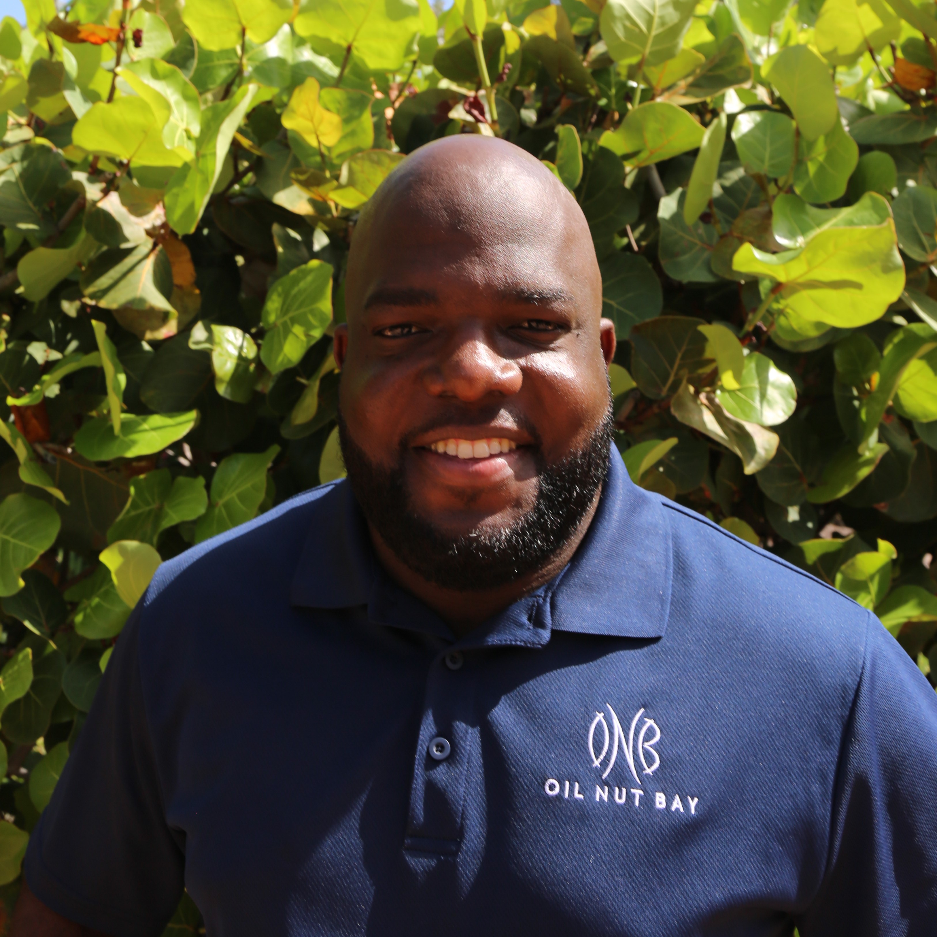 A smiling man with a bald head and beard is wearing a navy blue polo shirt with a white "Oil Nut Bay" logo. He stands in front of a lush green leafy background, embodying the charm that Dornelle Hazel often captures in her natural photography.