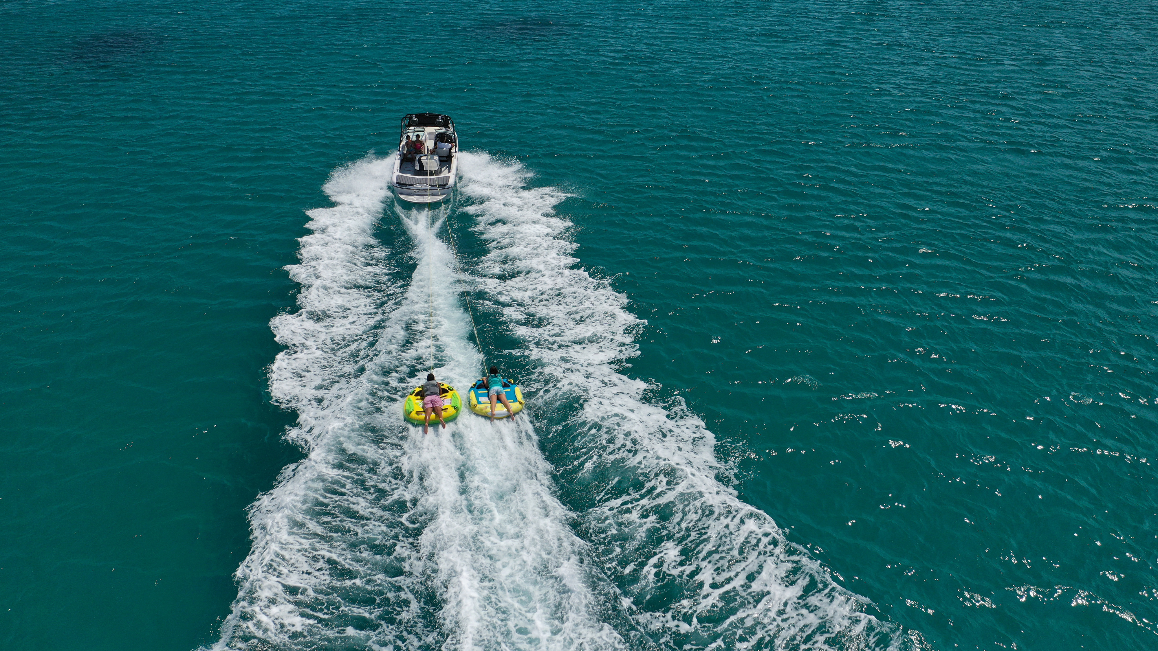 A motorboat pulls two people tubing on inflatable tubes across clear, turquoise water. The boat's wake creates white foam trails behind it as the tubers hold on and are towed across the sea. The sky is clear and the water is calm, suggesting a sunny day.