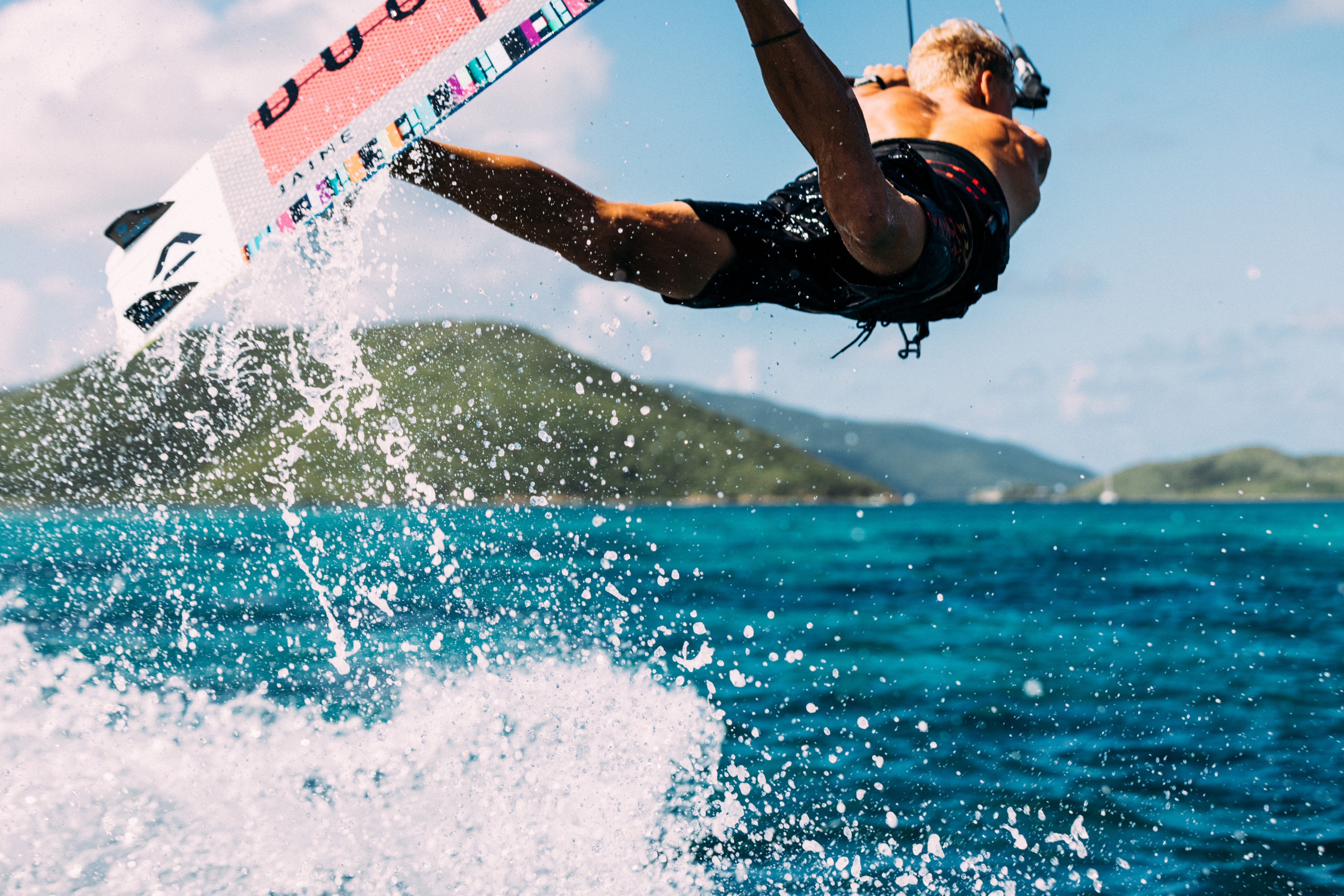 A person in mid-air performing an extreme kitesurfing trick above the ocean. The vibrant blue water splashes below, and green hills are visible in the background under a clear sky. The person is wearing shorts and holding onto the kite bar, epitomizing the thrill of water sports.