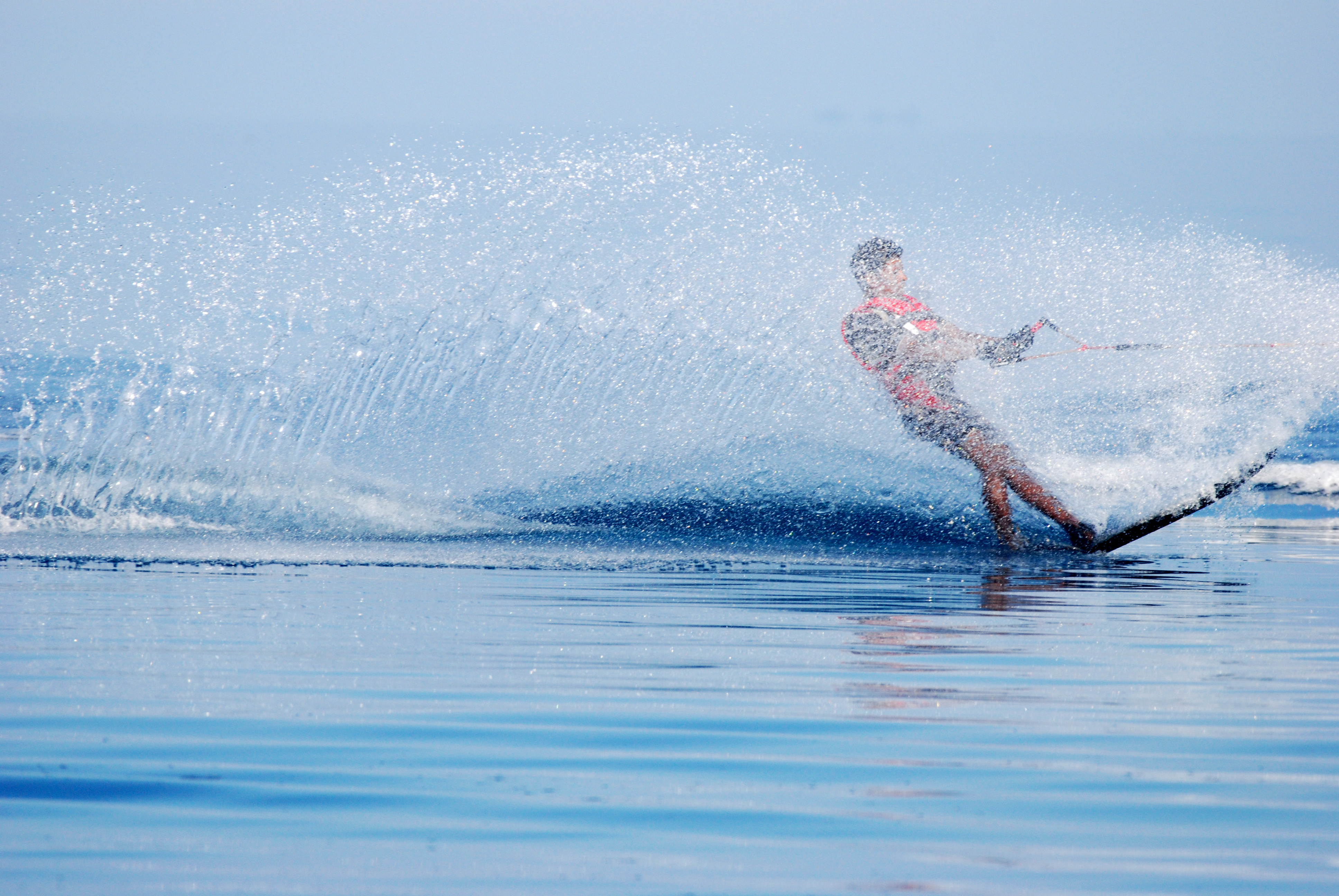 A person wearing a pink life jacket is waterskiing on a blue water surface, creating a large splash as they turn. Engaged in this exhilarating water sport, the skier holds onto a tow rope and is partially obscured by the arc of water spray. The sky is clear and blue.