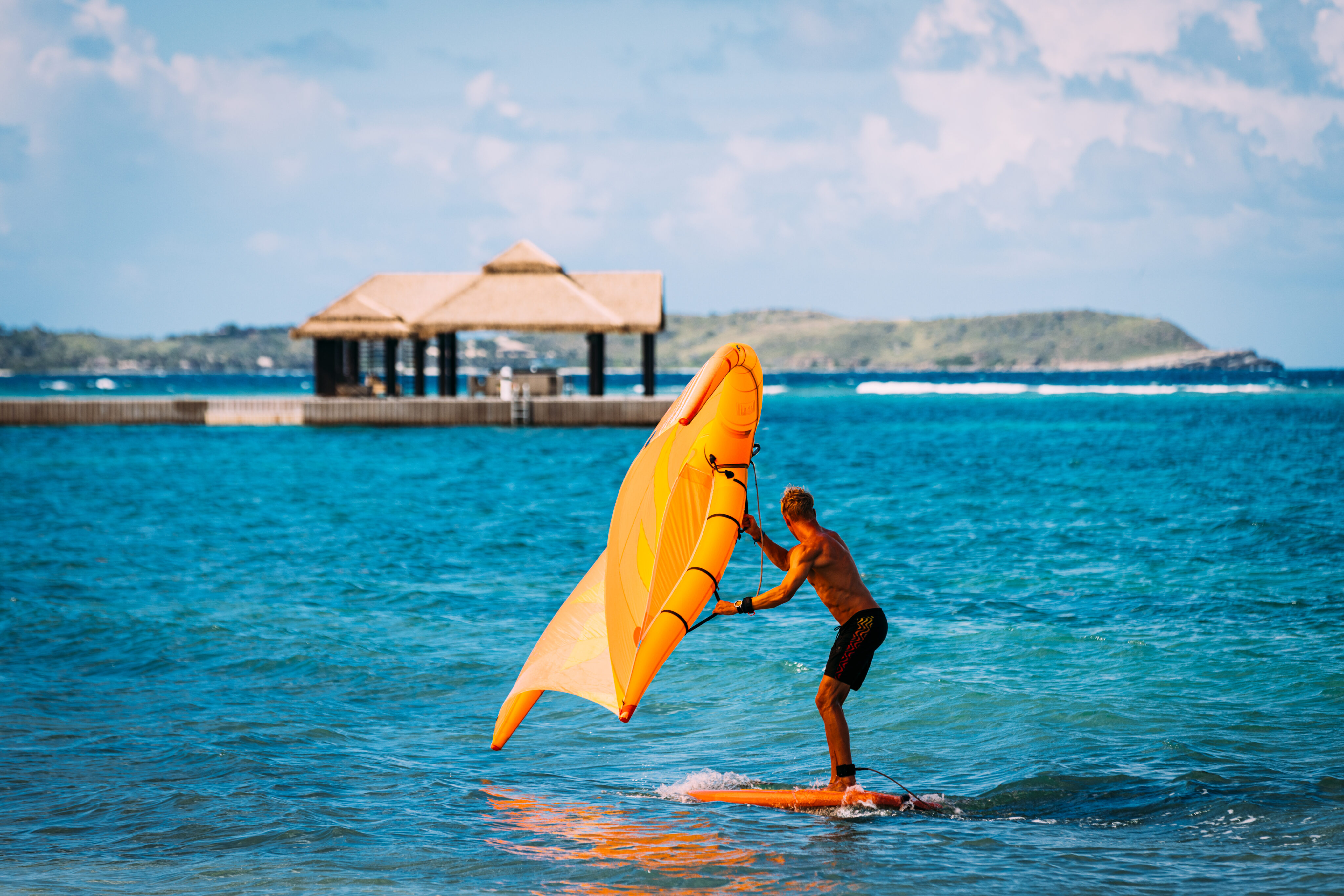 A person stands on a surfboard, engaging in wing surfing while holding an orange sail, gliding over the blue ocean. In the background, there is a wooden pavilion on stilts, with a scenic view of distant hills and a partly cloudy sky above.