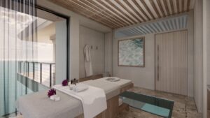 A serene massage room in a spa with a cushioned massage table next to glass doors leading to a balcony overlooking the ocean. The room has a neutral color palette, with a robe hanging on the wall, a decorative overwater-themed picture, and orchids in a glass vase.