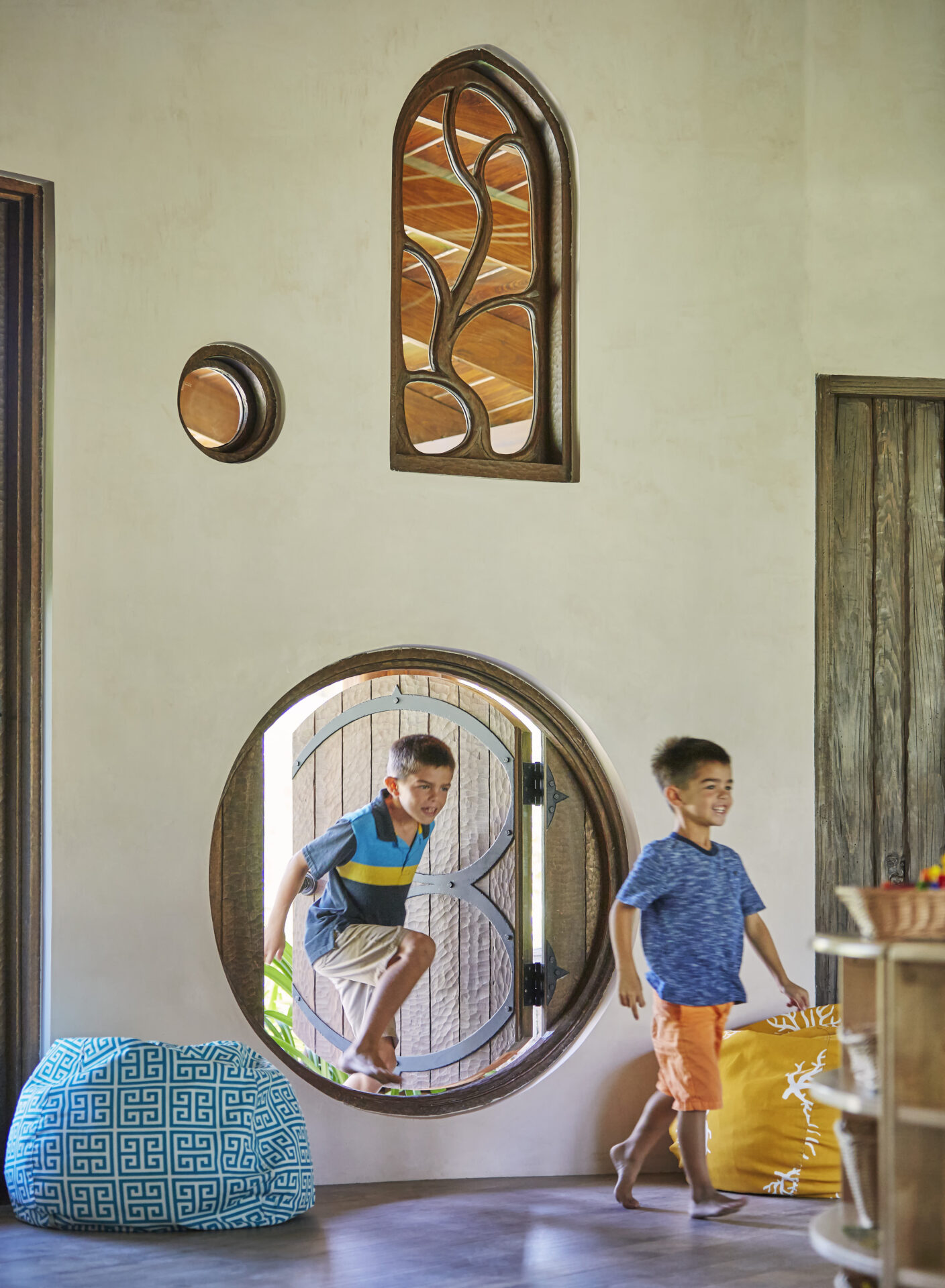 Two children playfully move through a circular, Hobbit-style door set in a light-colored wall with wooden accents. Decorative windows and round wall art adorn the space of this whimsical Kids Club. Large, colorful cushions are placed nearby, adding a cozy, playful atmosphere to the room.