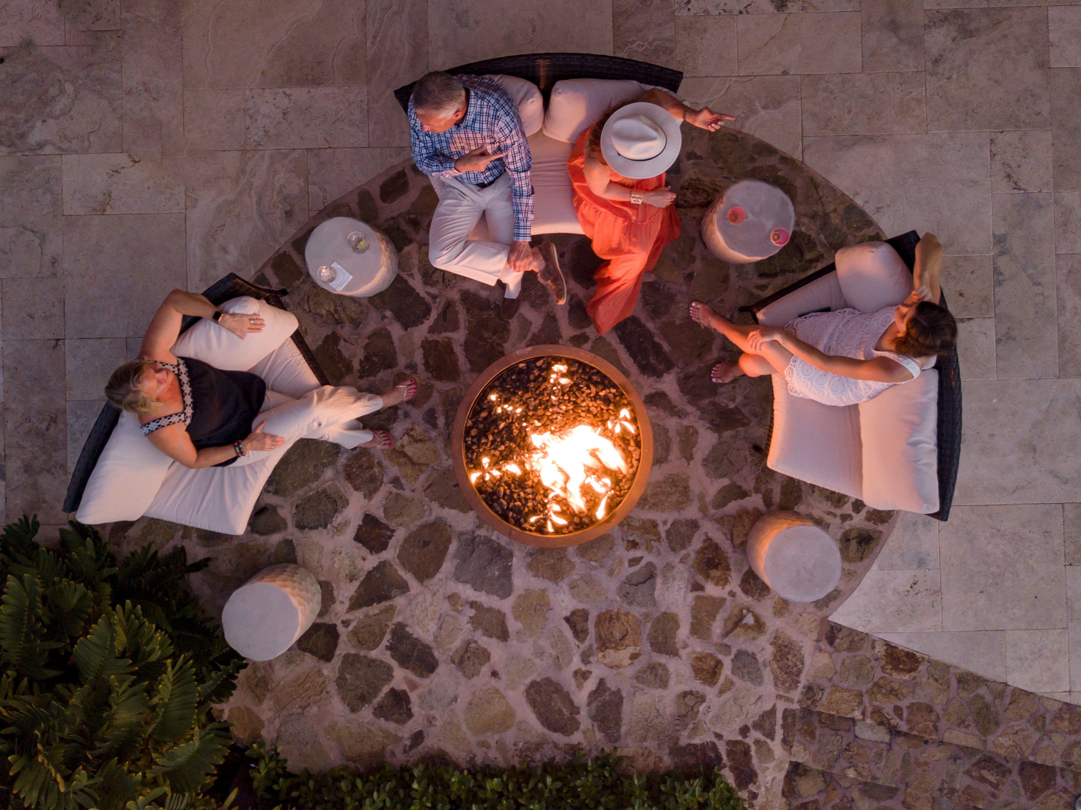 Overhead view of four people sitting around a circular fire pit on a stone patio. They are seated on cushioned outdoor furniture, engaged in conversation about life in the British Virgin Islands. Drinks are placed on small tables around them. The surroundings are made of stone and tiled flooring.