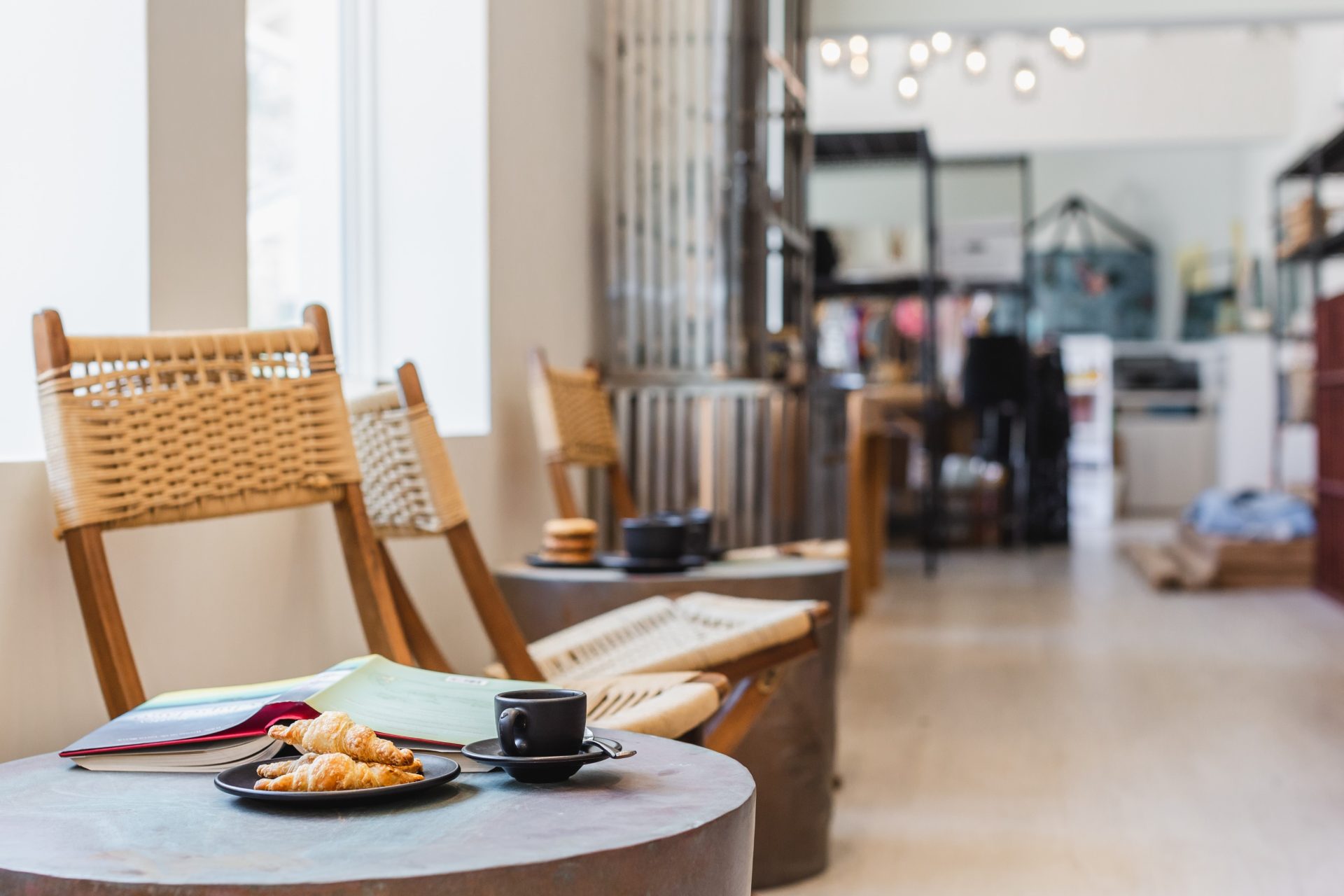 A cozy marina café interior featuring wooden chairs with woven backs along a light-filled window. A small table in the foreground holds a croissant on a plate, an open book, and a black coffee cup. The background has blurred shelves and hanging lights.