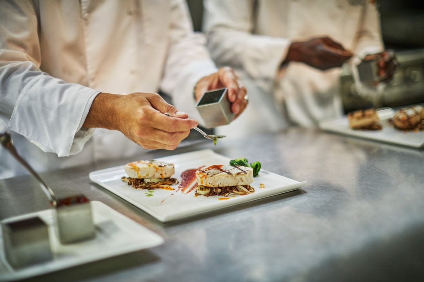 Chefs in white uniforms carefully plating gourmet dishes on a stainless steel surface. One chef is using a small spoon to add garnish to a square white plate, while the others work in the background preparing similar dishes for an exquisite In-Villa Dining experience.