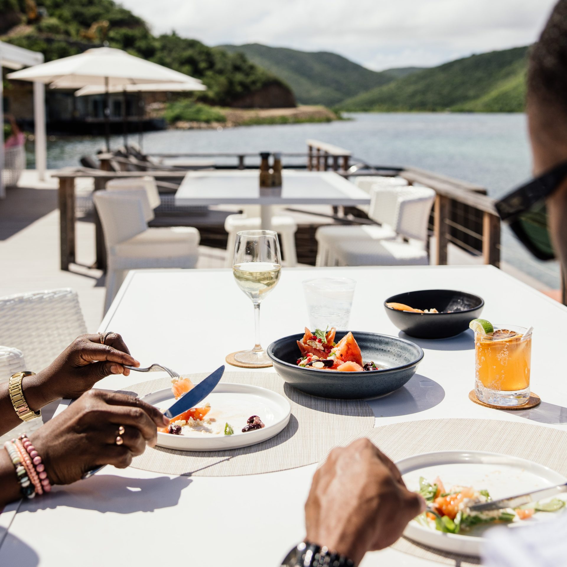 Two people enjoying a waterfront dining experience at the scenic Nova restaurant, with hills and a shimmering body of water as their backdrop. Their table boasts plates of food, a glass of white wine, a cocktail, and water. The background includes a deck with more seating and umbrellas.