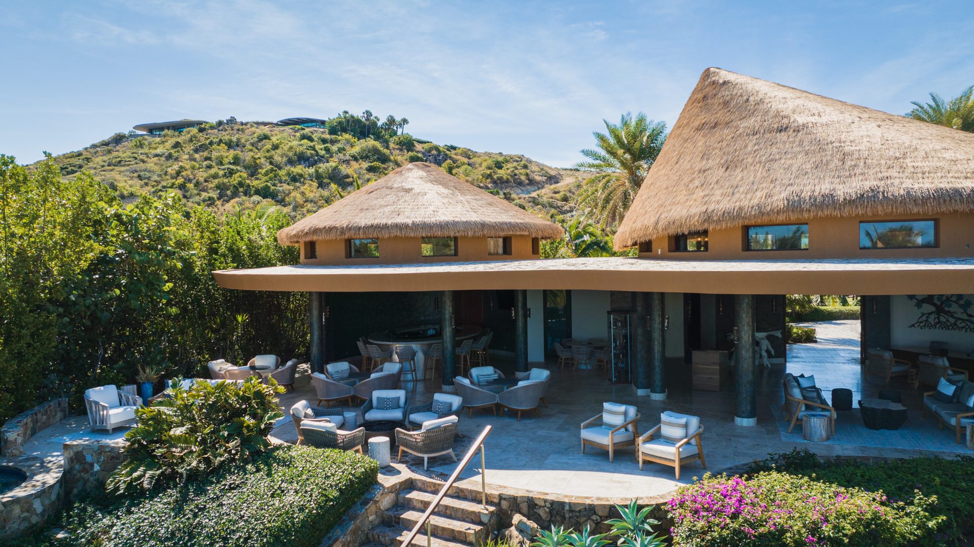 A luxurious tropical resort features thatched-roof structures and a Pavilion Lounge with open-air seating areas, complete with comfortable chairs and tables amidst lush greenery. Stone steps lead to the main lounging area. Hills and trees embrace the tranquil setting under a clear blue sky.