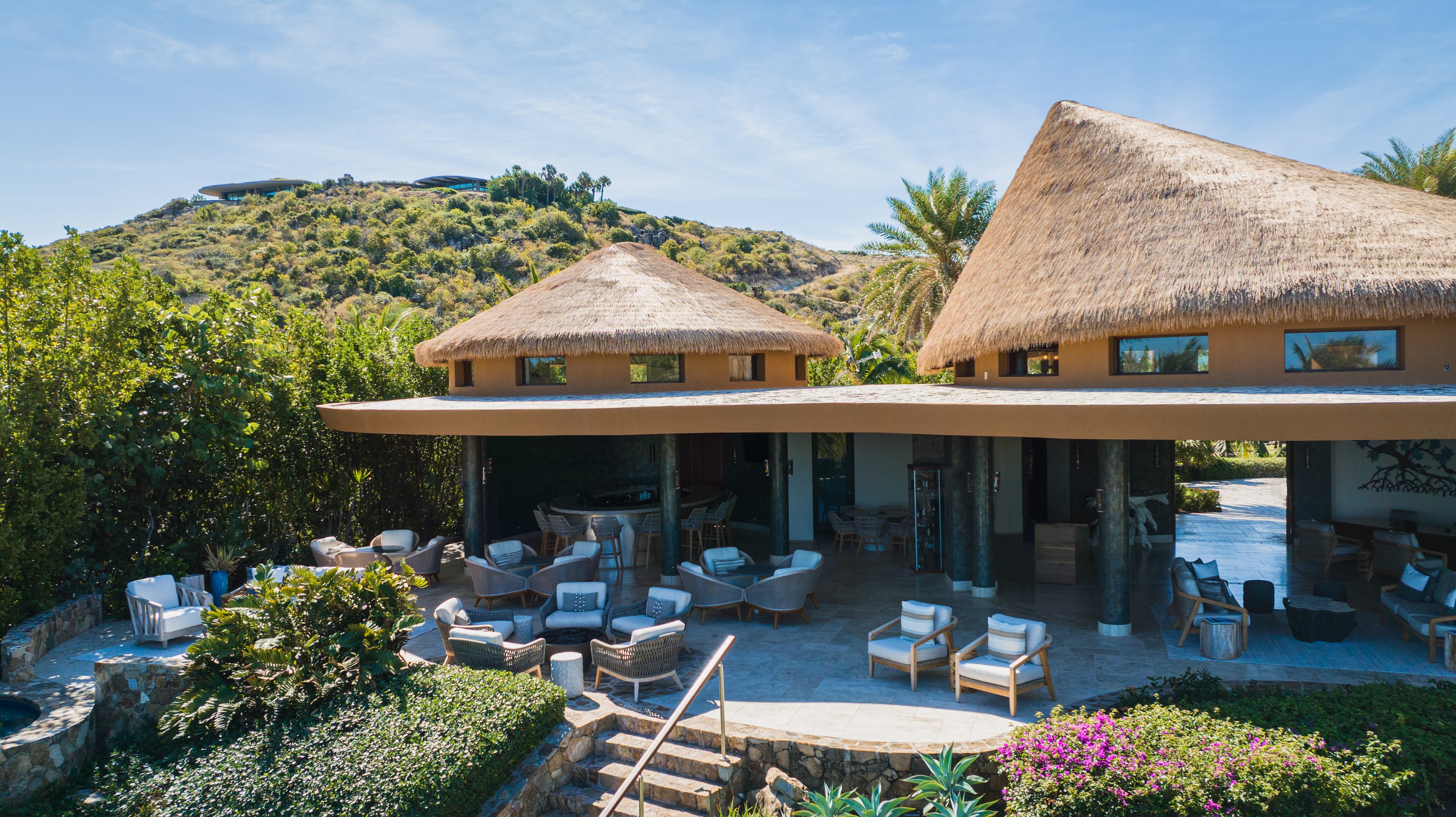 A luxurious tropical resort features thatched-roof structures and a Pavilion Lounge with open-air seating areas, complete with comfortable chairs and tables amidst lush greenery. Stone steps lead to the main lounging area. Hills and trees embrace the tranquil setting under a clear blue sky.